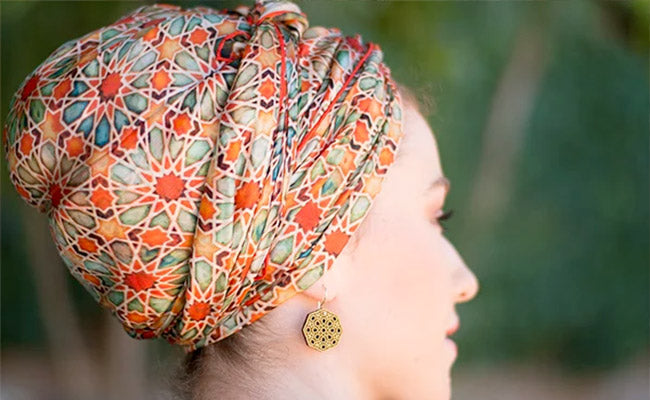 A woman in profile wears a colorful patterned headwrap with geometric designs and a matching dangling earring. The background is blurred and outdoors.