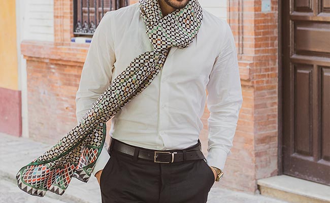 Man wearing a white shirt, black pants, and a large patterned scarf, walking outdoors by a brick and white building with his hands in his pockets.