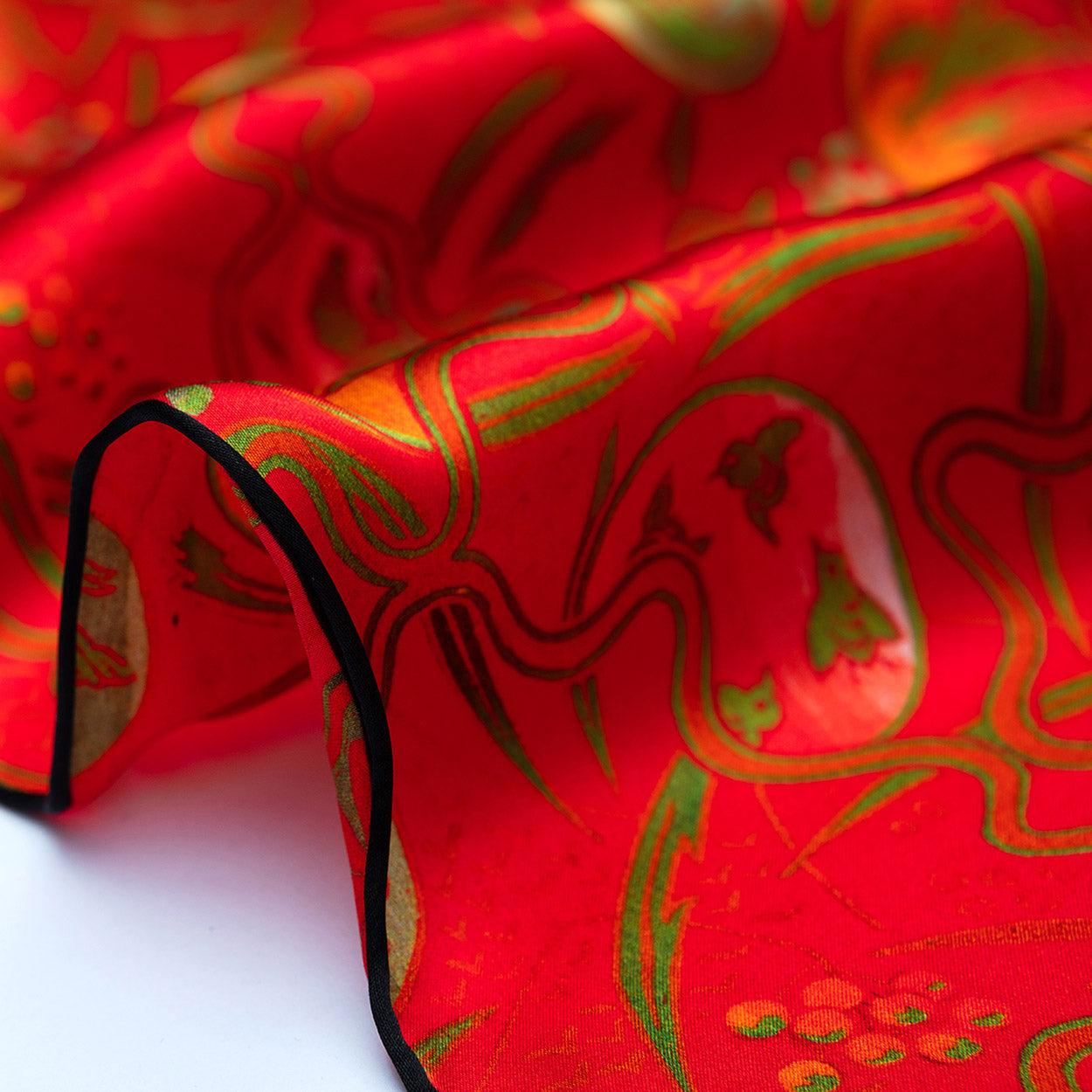 A close-up of the Red Silk Scarf with Abstract Floral Print Kabuki, showing red and black fabric with gold and green patterns, detailed with a black trim along the edge, draped in soft folds.