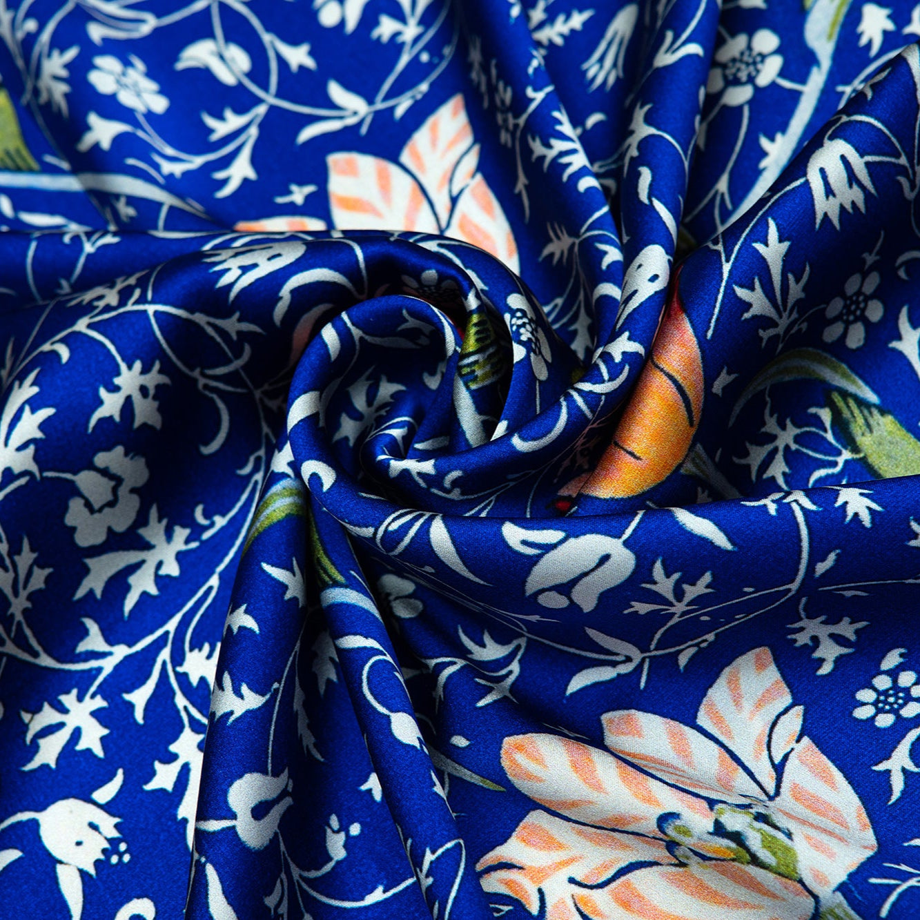 The Floral Blue Silk Scarf Galata features a smooth, silky texture with Art Nouveau-inspired orange, green, and white floral and bird patterns on vibrant blue, gathered and twisted at the center for a luxurious touch.
