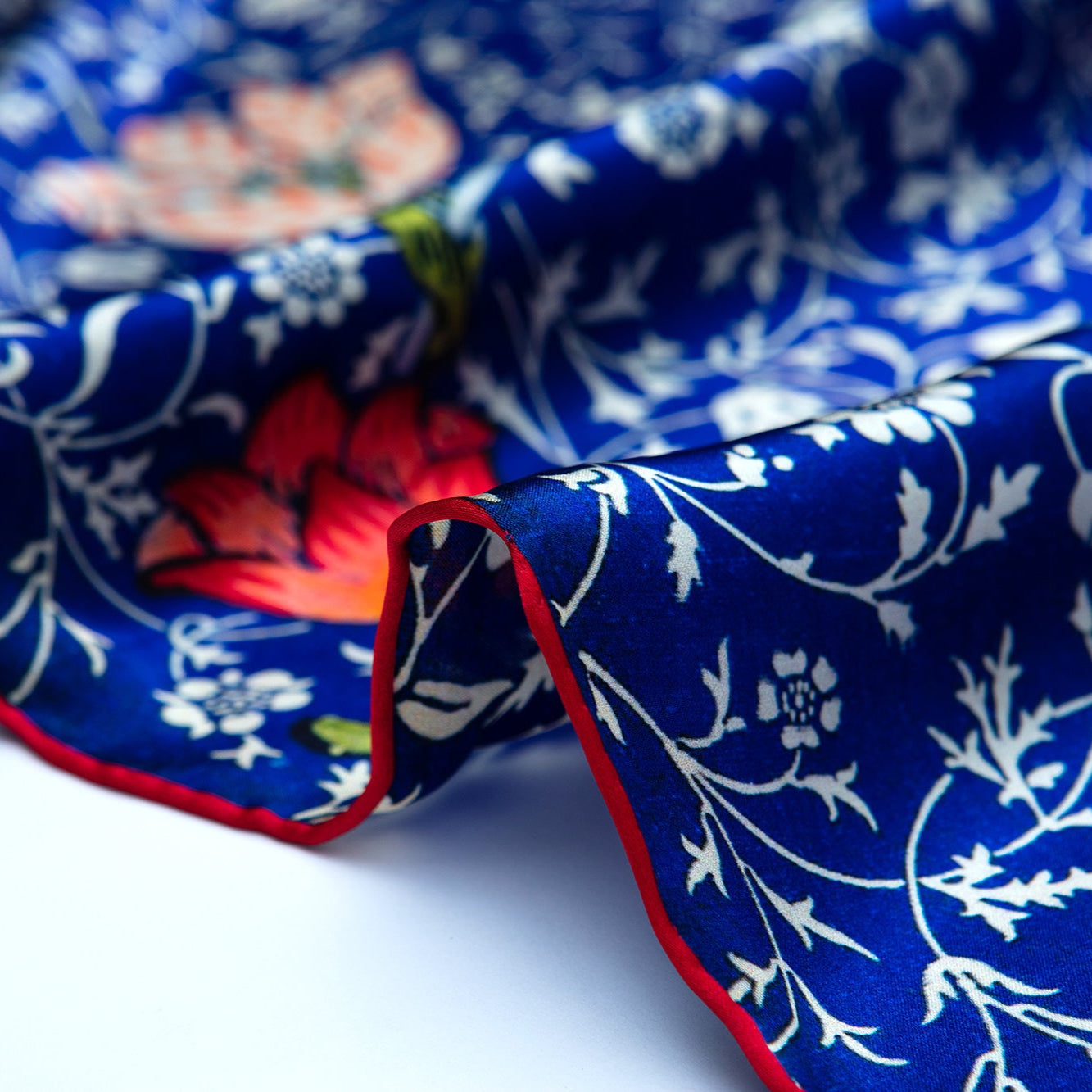 A close-up of the Floral Blue Silk Scarf Galata: luxurious blue silk with white floral patterns, bold red and pink Art Nouveau flowers, and a red-edged hem, gently draped on a white surface.