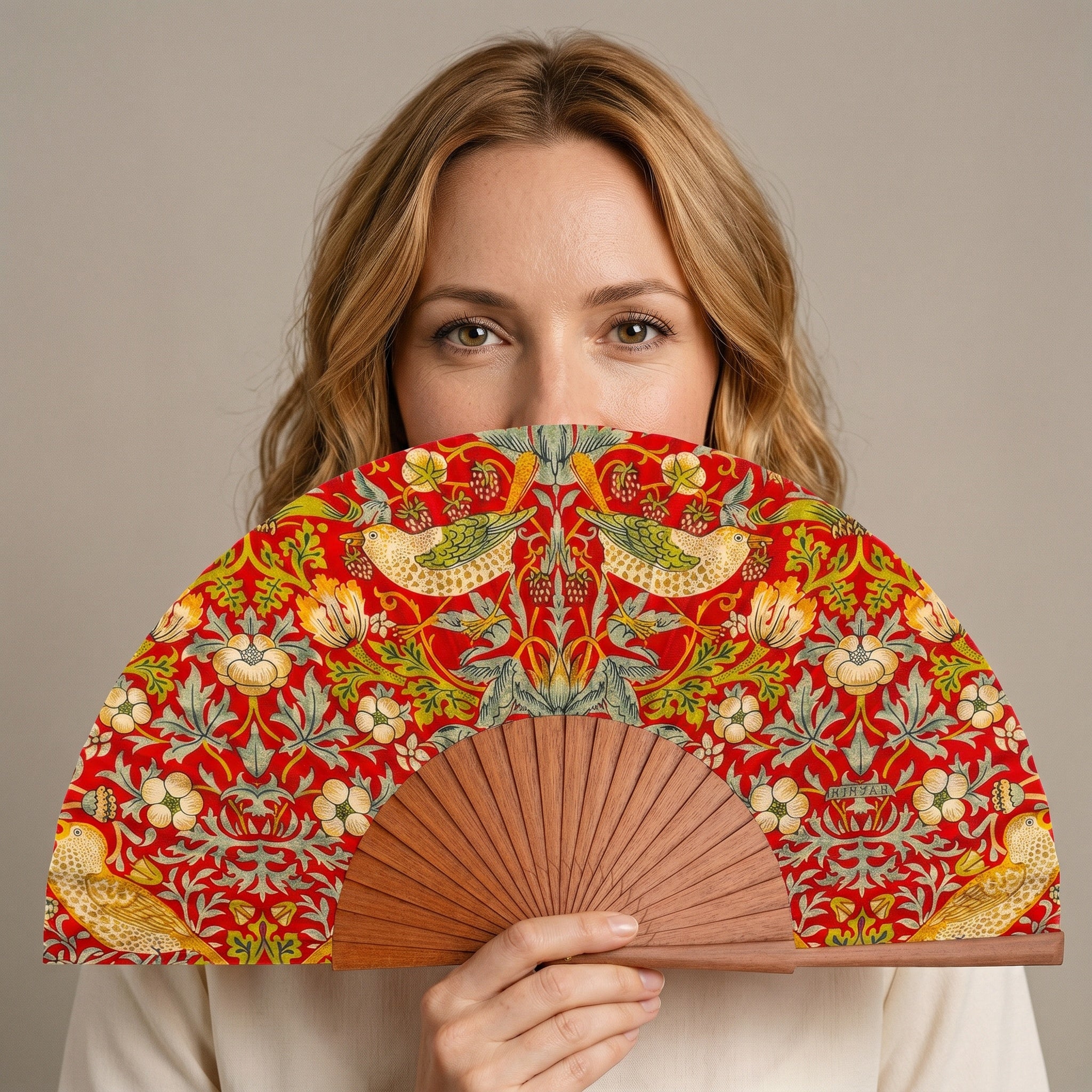 A woman holding the William Morris Print Hand Fan Birds, with its William Morris Strawberry Thief print, against a soft neutral background.