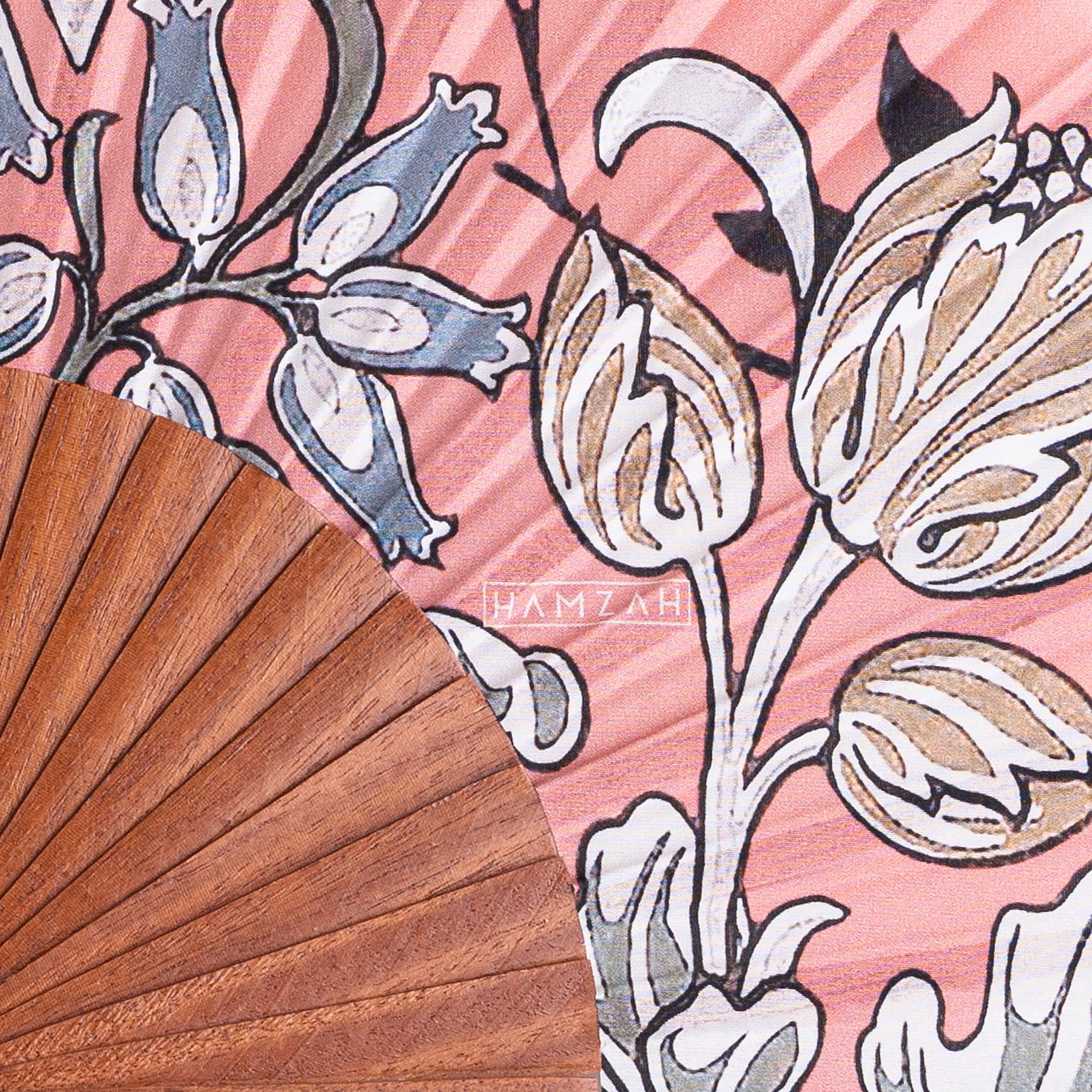 A close-up of the Folding Silk Fan with Floral Print Elinor partially covers a pink fabric decorated with white and brown florals, inspired by Spanish craftsmanship, with the name HAMZAH printed in white on the silk.