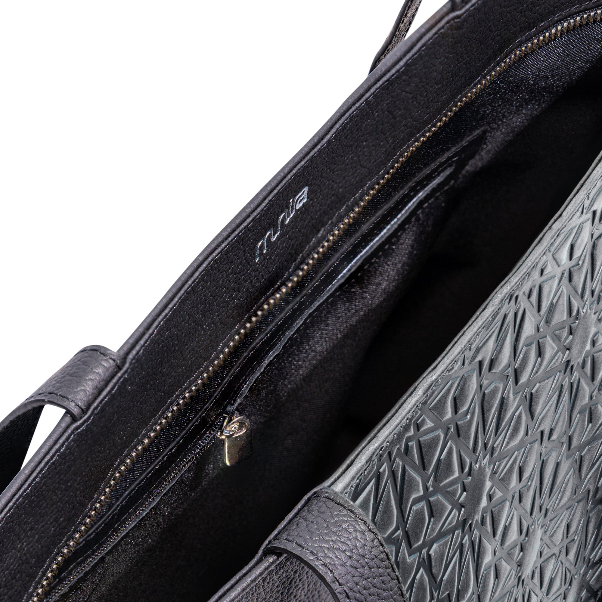 Close-up of the Black Leather Tote Bag - Alhambra Pattern Embossed, featuring a textured geometric exterior, a zippered interior pocket, and the MUA logo printed inside.
