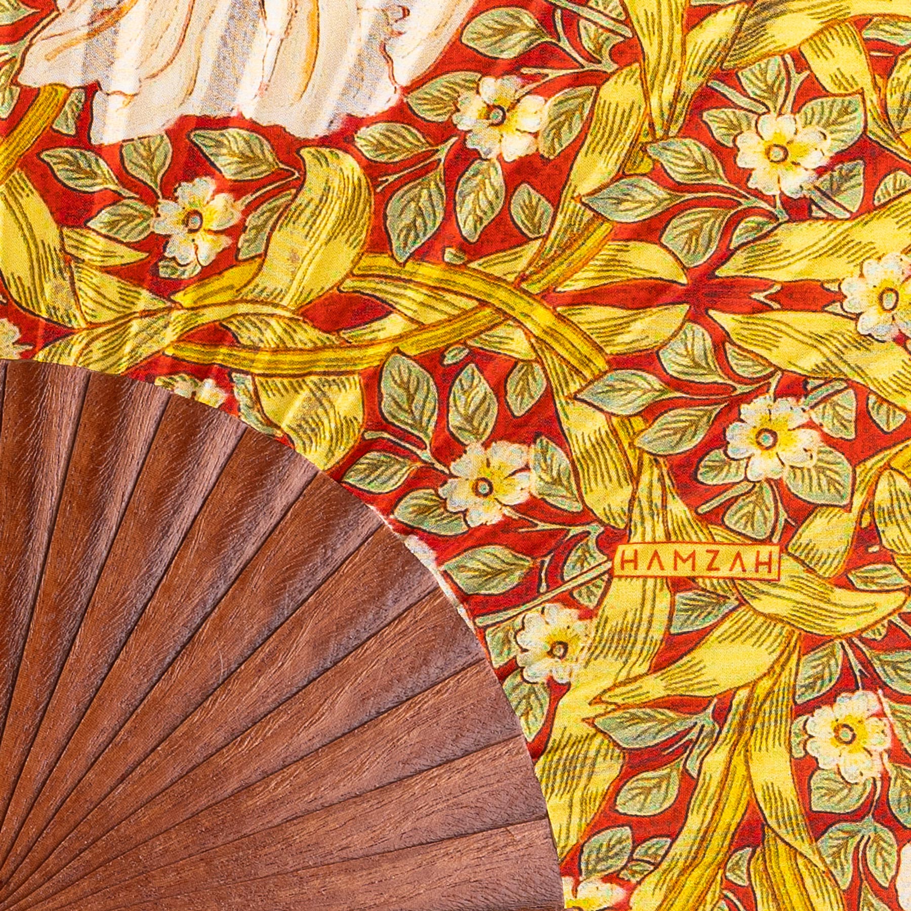 A close-up of the Hand Fan Made in Spain Basile: its top displays a floral silk pattern with white flowers, green leaves, and yellow ribbons on red, while the lower section is brown wood. The fan features "Hamzah" on the fabric.