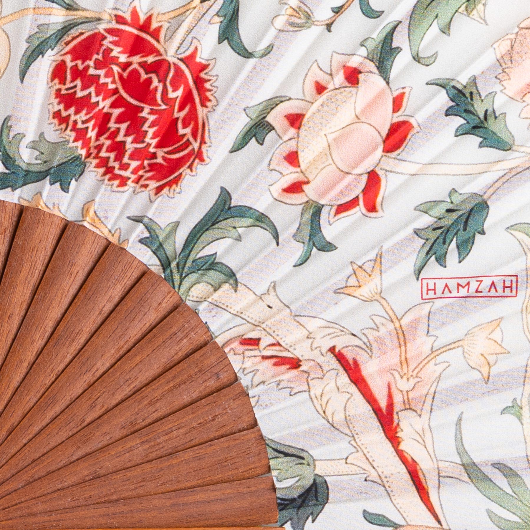A close-up of the Floral Design Fan William Morris Print Odette, featuring a wooden base and fabric with red and cream floral patterns, green leaves, and "Hamzah" in red text, evoking Spanish craftsmanship.