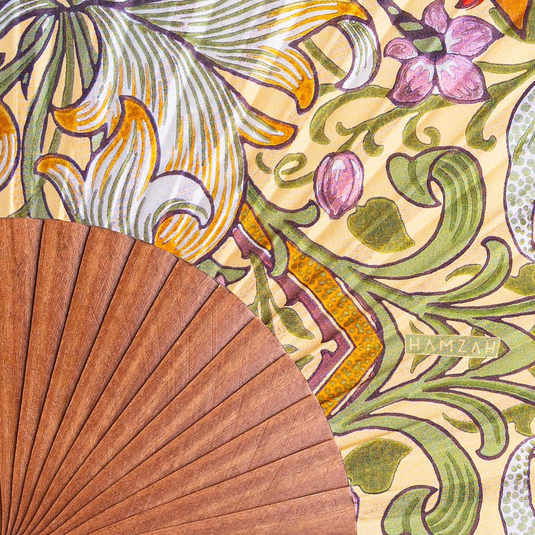 A Spanish Hand Fan with Floral Print Georg lies on vibrant floral fabric featuring green, yellow, and pink designs. The name HAMZAH appears on the silk material.