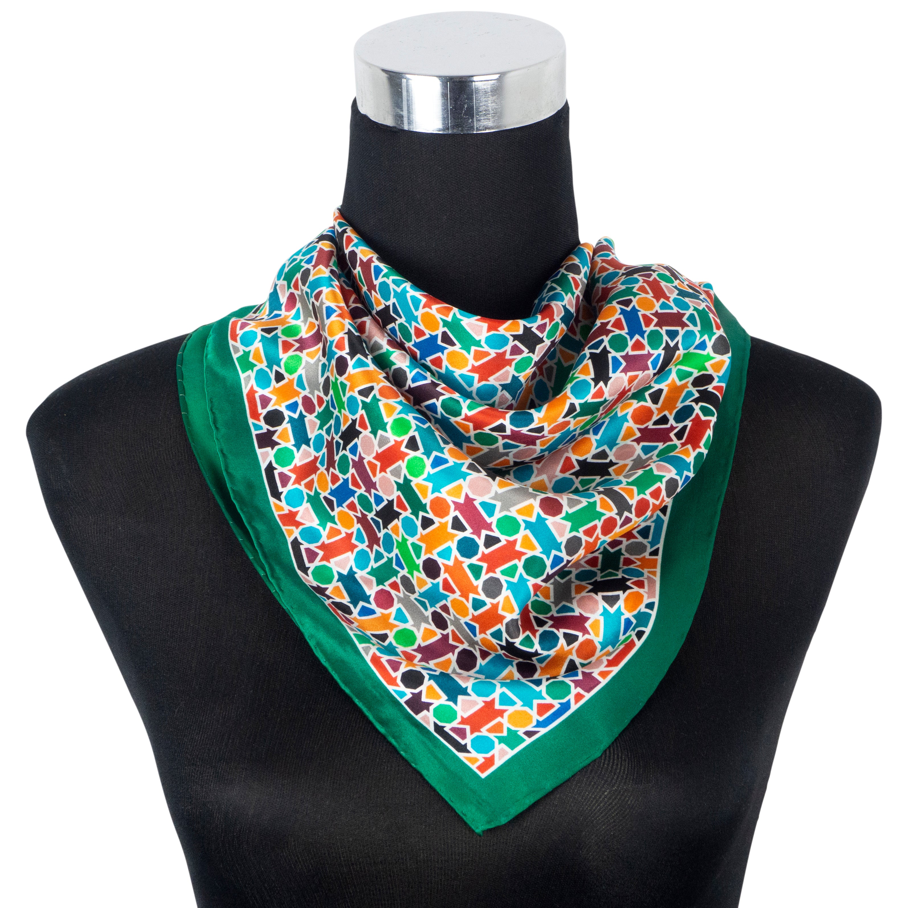 A mannequin displays the Multicolor Silk Square Scarf Amal, draped in a triangle around the neck. Made of silk with hand rolled hems and a green border, it features a vibrant abstract geometric pattern.