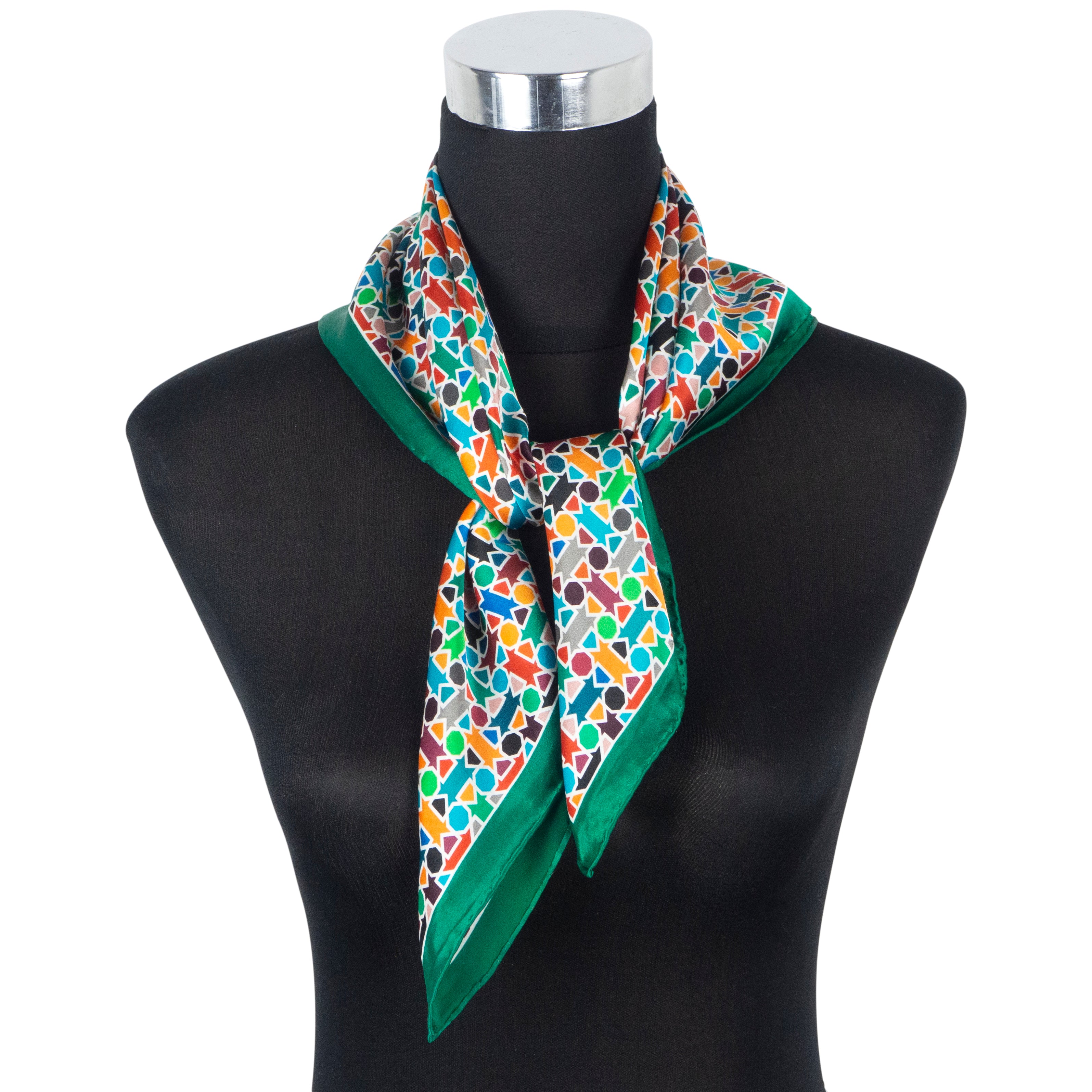 The Multicolor Silk Square Scarf Amal, featuring hand rolled hems and a geometric pattern with green borders, is tied around the neck of a black dress form mannequin topped with a silver cap.