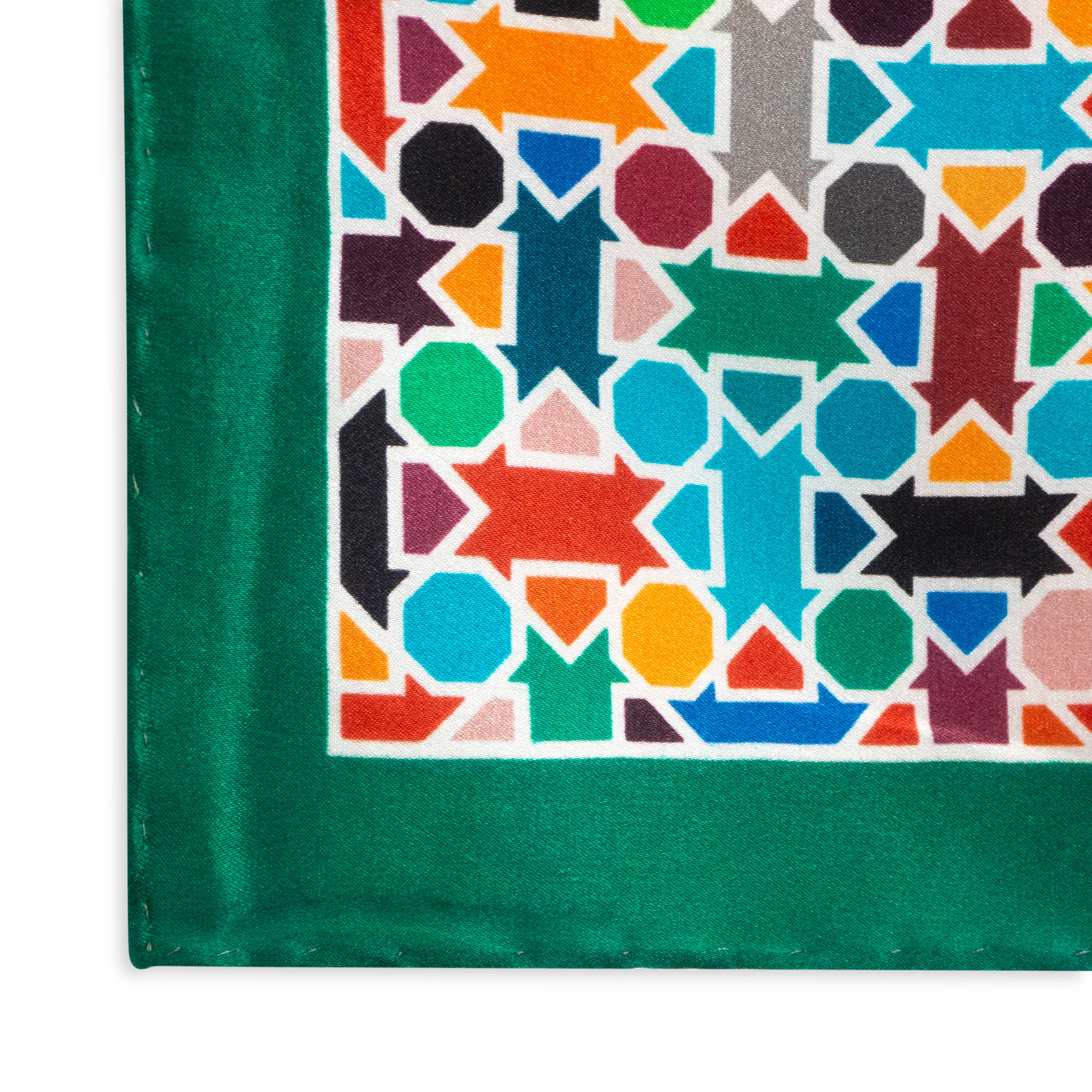 The Multicolor Silk Square Scarf Amal features a vibrant geometric pattern of arrows and hexagons in red, blue, orange, green, and black on white silk, finished with a bright green satin border and hand rolled hems.