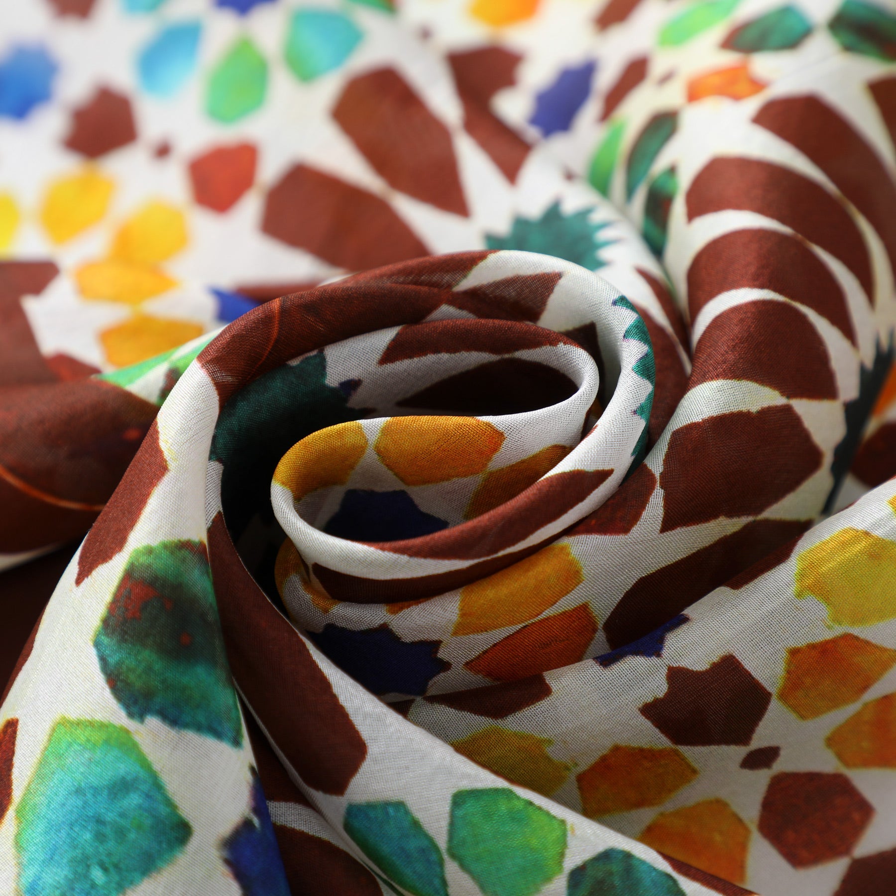 A close-up of the Brown and Yellow Silk Scarf Garnata, rolled in a spiral, displays its soft texture and vibrant geometric mosaic patterns inspired by Islamic Art in red, yellow, green, and blue tones.