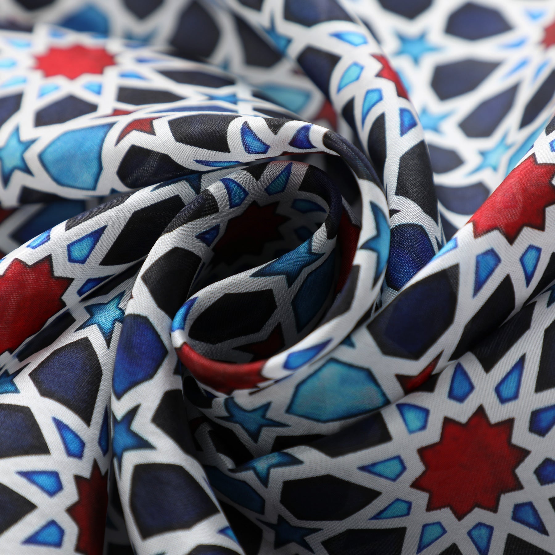 The Alhambra Inspired Silk Scarf Fatima features a swirling close-up of intricate red, blue, black, and white geometric star patterns. The softly gathered silk highlights its vibrant colors and luxurious texture—a timeless accessory.