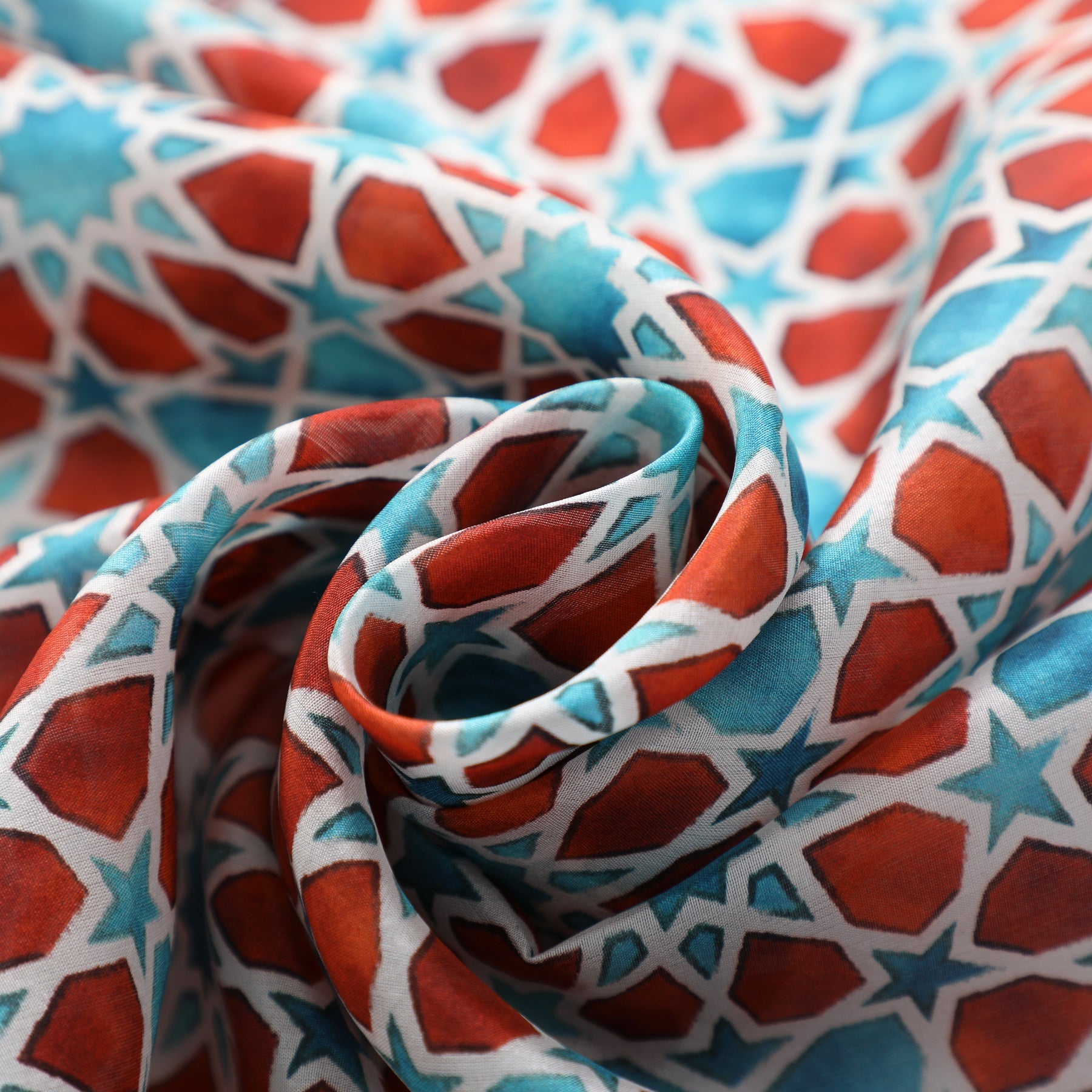A close-up of the Brown and Blue Silk Scarf Fatima, folded in a loose spiral to highlight its blue geometric patterns inspired by Al-Andalus, with soft curves and shadows.