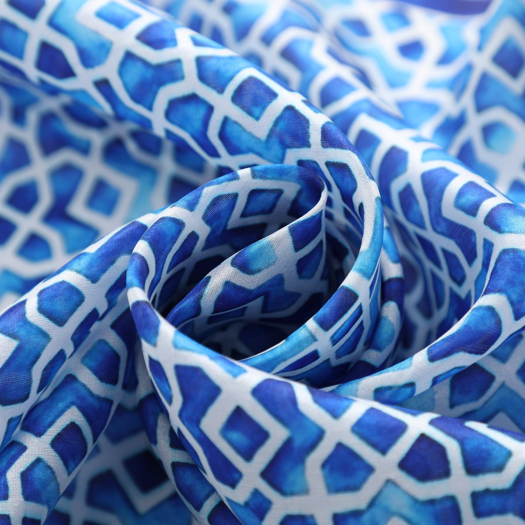 Close-up of the Oblong Blue Silk Scarf Alcazaba, softly folded to highlight its blue and white geometric diamond pattern, with gentle curves inspired by Islamic art.