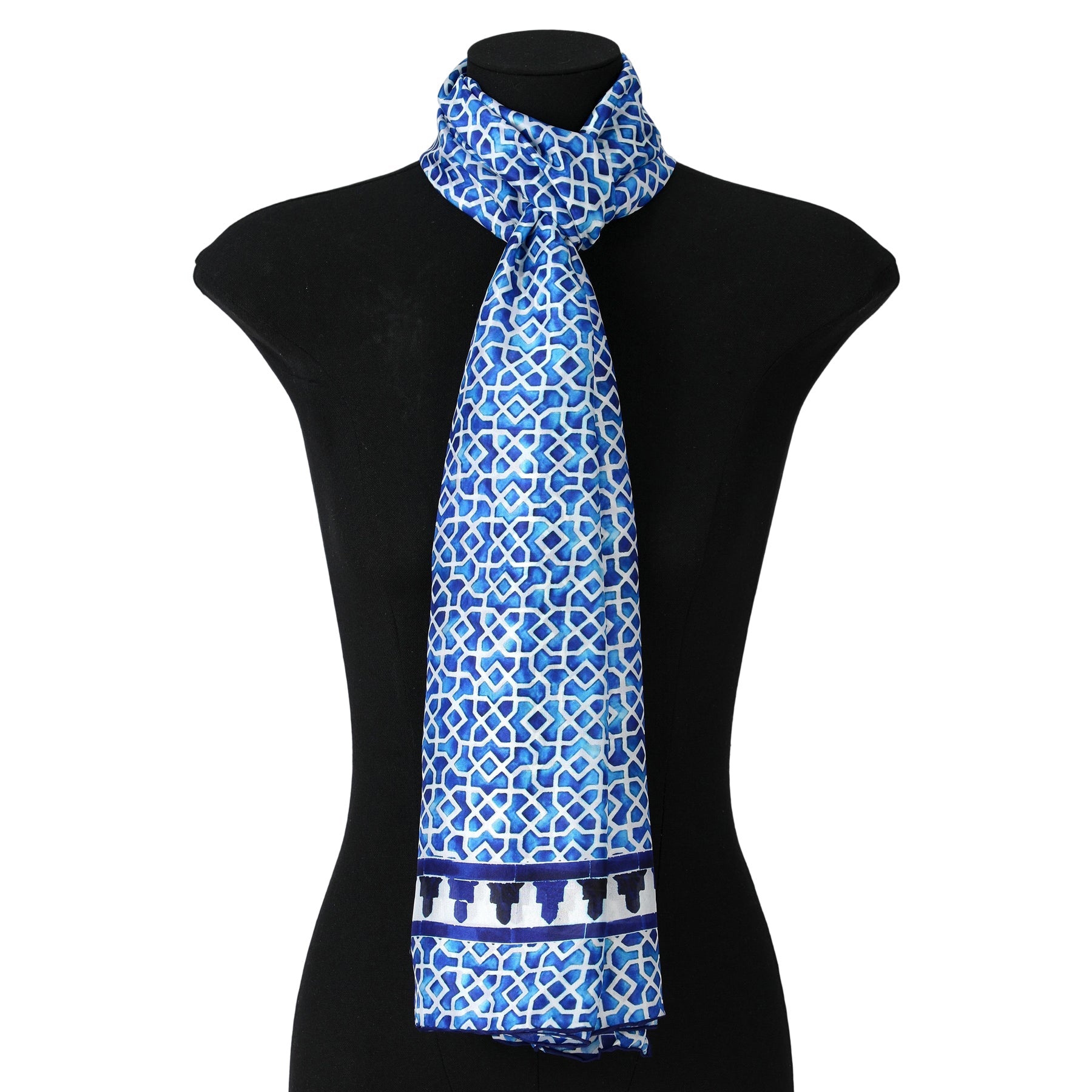 A mannequin displays the Oblong Blue Silk Scarf Alcazaba, featuring blue and white geometric designs inspired by Islamic art and a decorative border, loosely tied around the neck.
