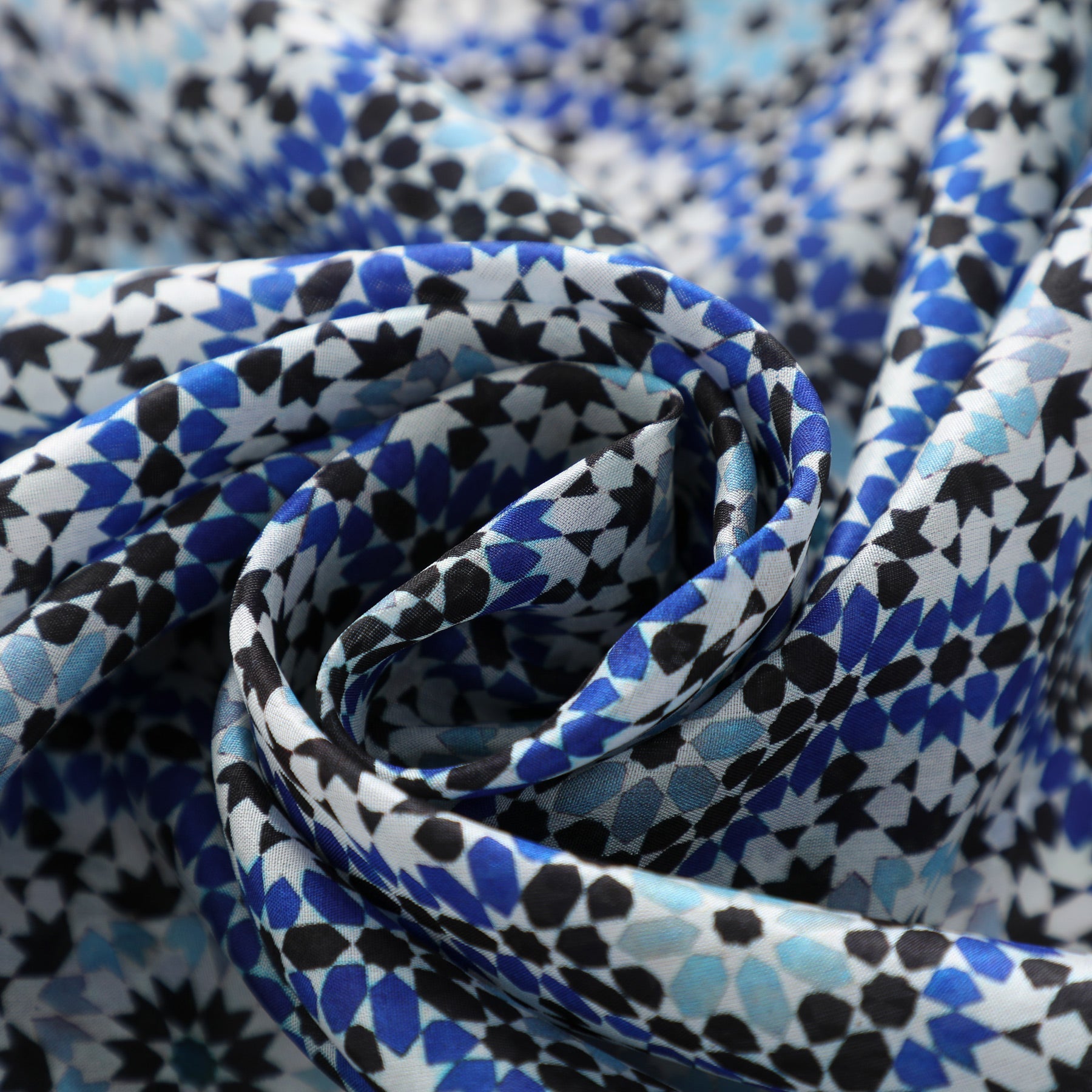 Close-up of the Moroccan Tiles Inspired Silk Scarf Masdar, showcasing a geometric mosaic pattern in blue, black, and white, arranged in a spiral fold.