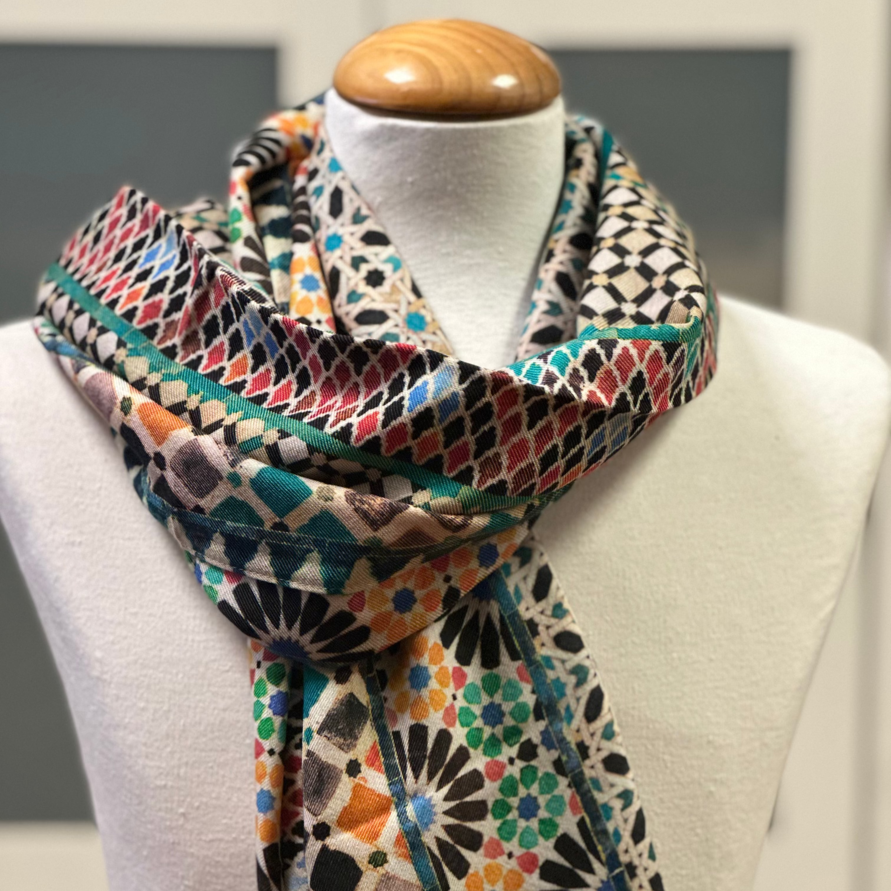 A mannequin displays the Silk and Wool Scarf Mehdya, featuring geometric and floral mosaic patterns inspired by Islamic art, draped elegantly in vibrant teal, orange, black, red, and beige.