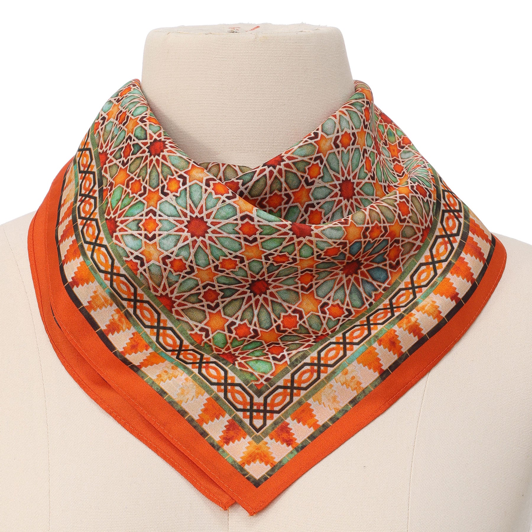A mannequin displays the Orange Square Scarf Zellige, featuring intricate Islamic-inspired geometric patterns in orange, green, red, and cream with a solid orange border, stylishly draped around the neck.