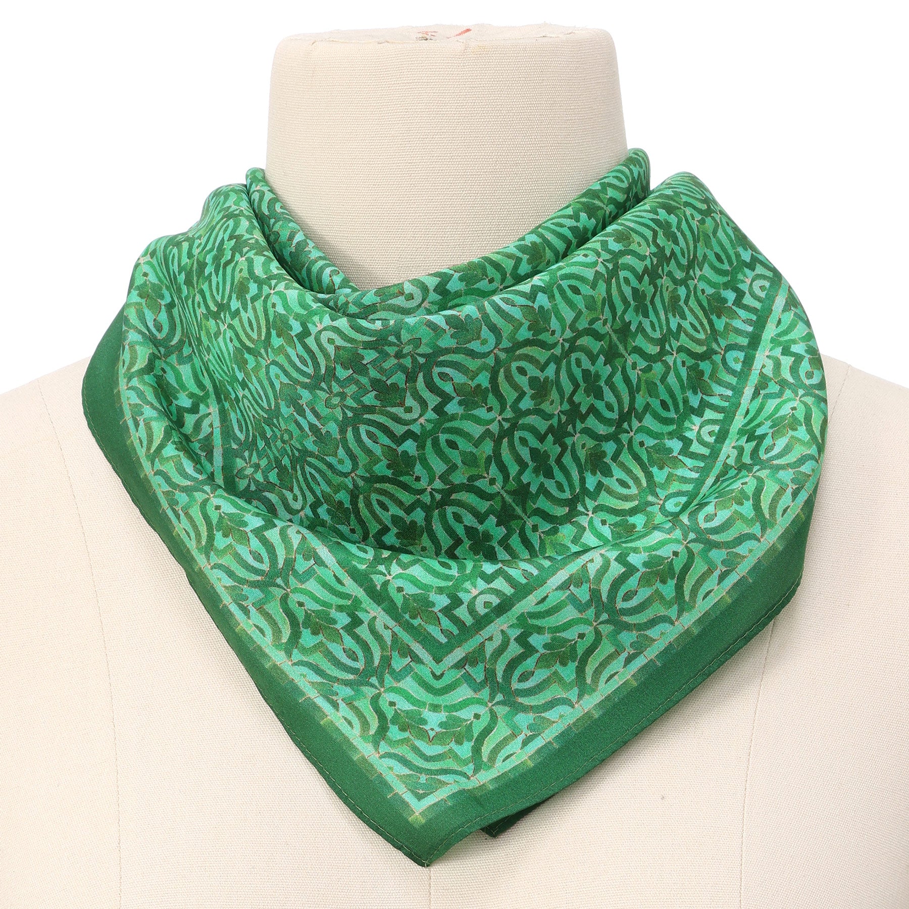 The Green Square Silk Scarf Fihri, featuring an intricate Islamic art-inspired design, is folded into a triangle and draped around the neck of a white mannequin bust.
