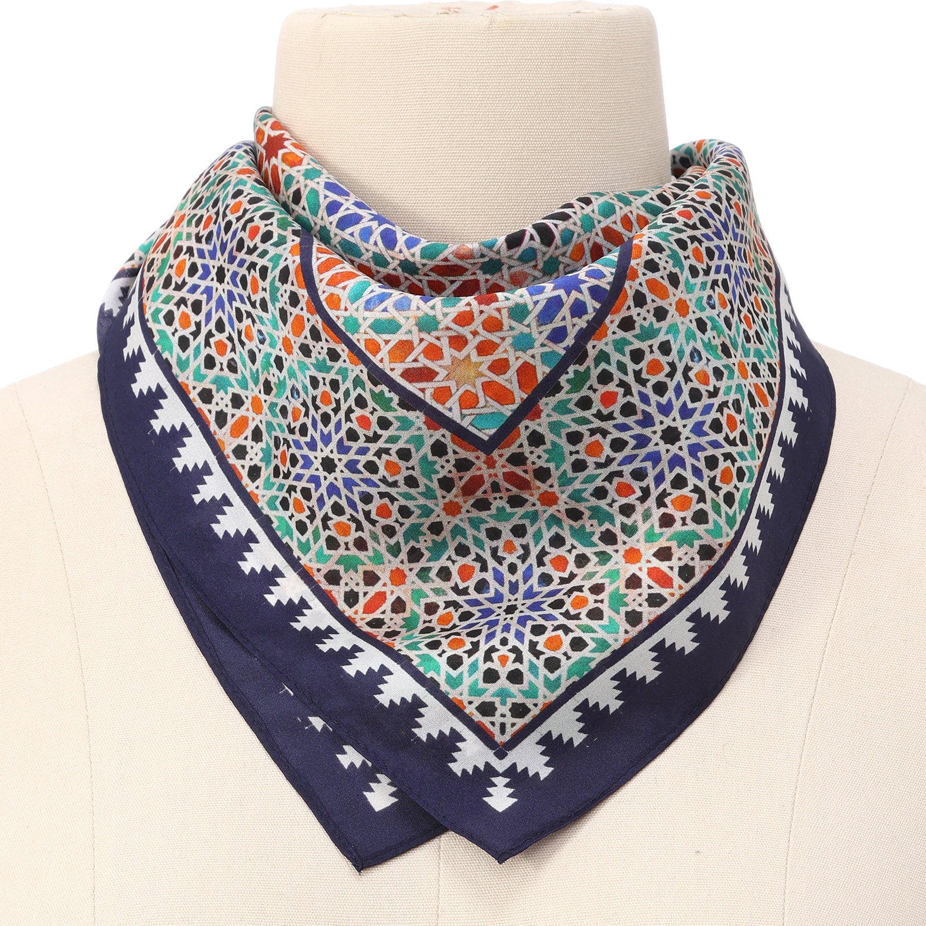 A mannequin showcases the Square Scarf Blue Madiya, featuring intricate geometric patterns in green, red, orange, and blue, bordered by a navy blue edge with a white zigzag design.
