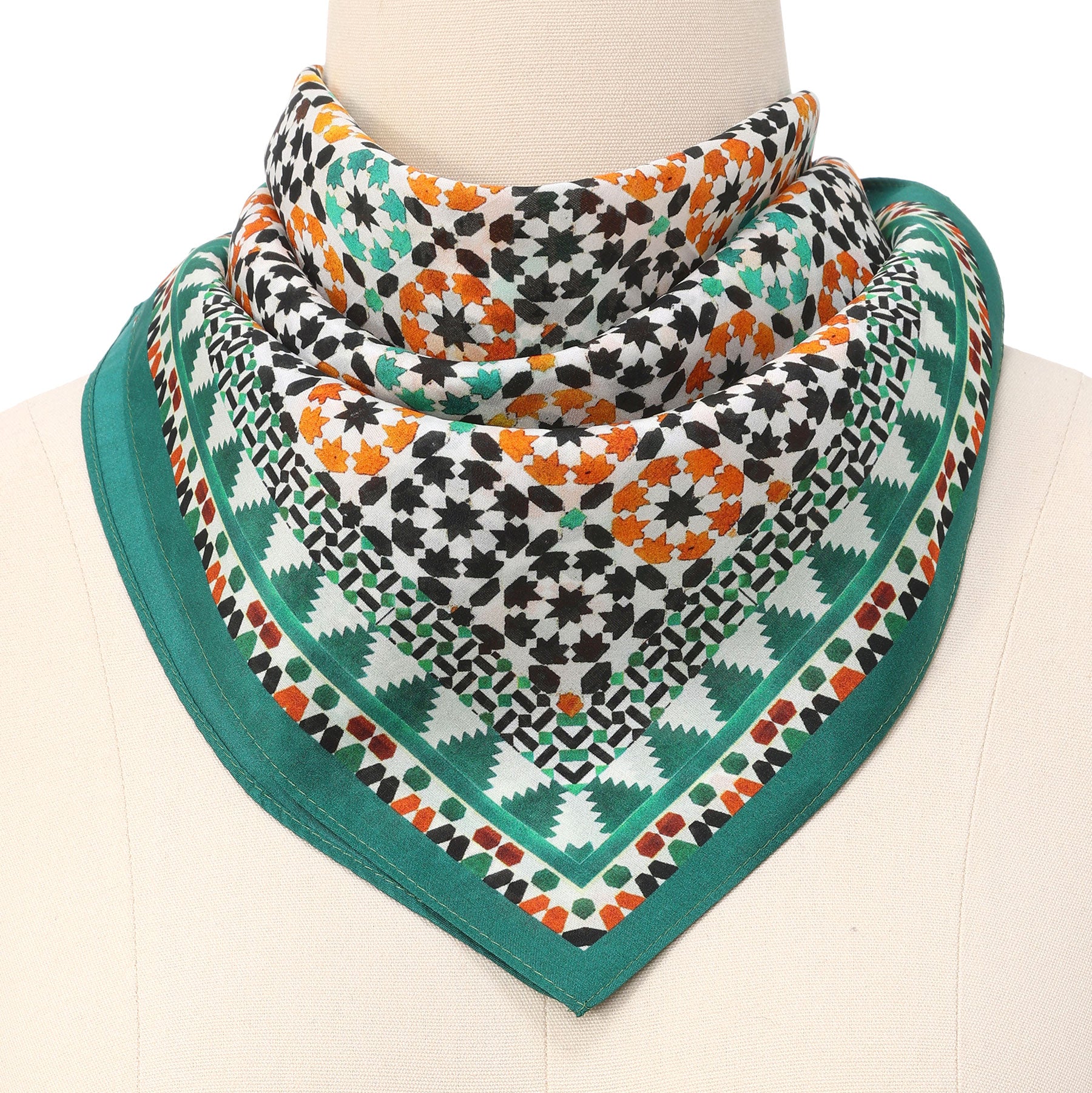 A mannequin showcases the Green Square Silk Scarf Sabika, featuring a geometric Islamic art-inspired print in green, orange, black, and white with triangles, diamonds, rosettes, and a solid green border.