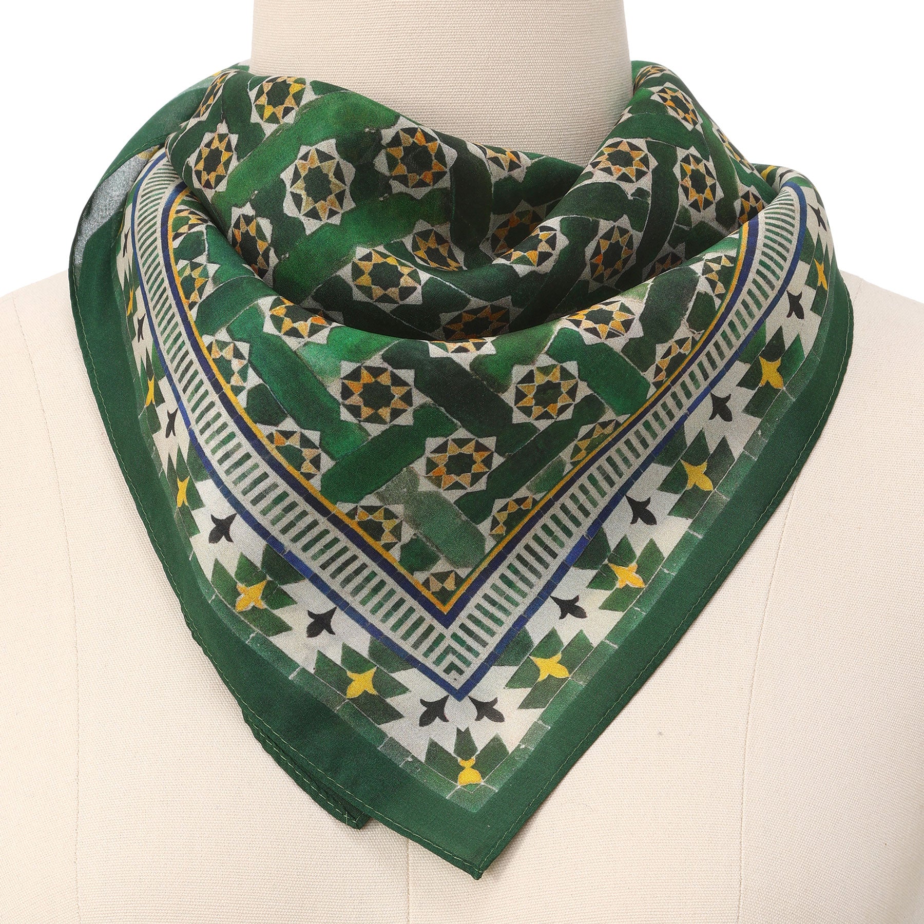 The Green Square Scarf Silk, featuring a geometric pattern of yellow, white, and green star shapes inspired by Islamic art, is folded into a triangle and wrapped elegantly around a mannequin’s neck.