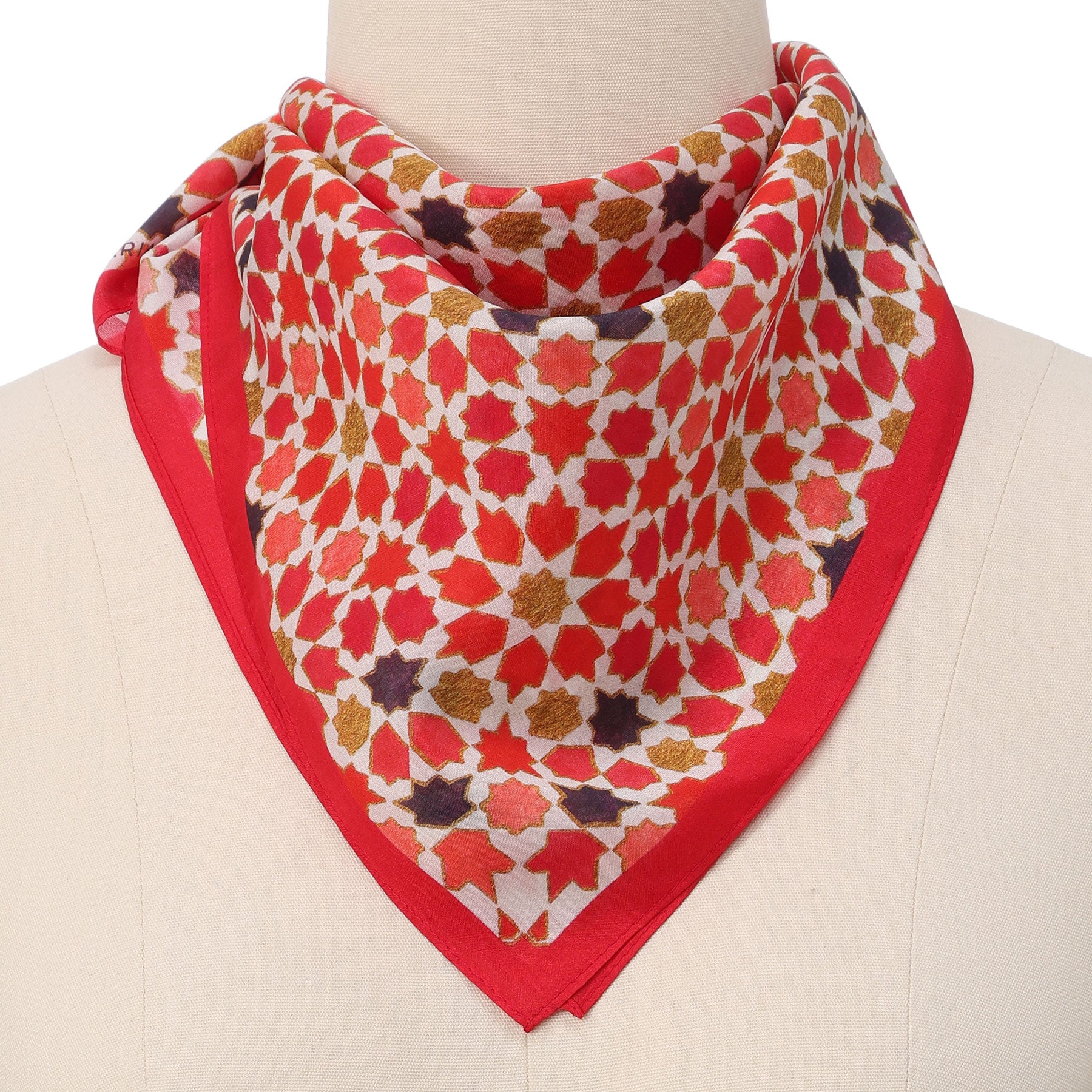 A mannequin displays the Small Square Scarf Alhambra—silk, with red, gold, and purple star patterns inspired by Islamic art—folded and draped around the neck, bordered in solid red.