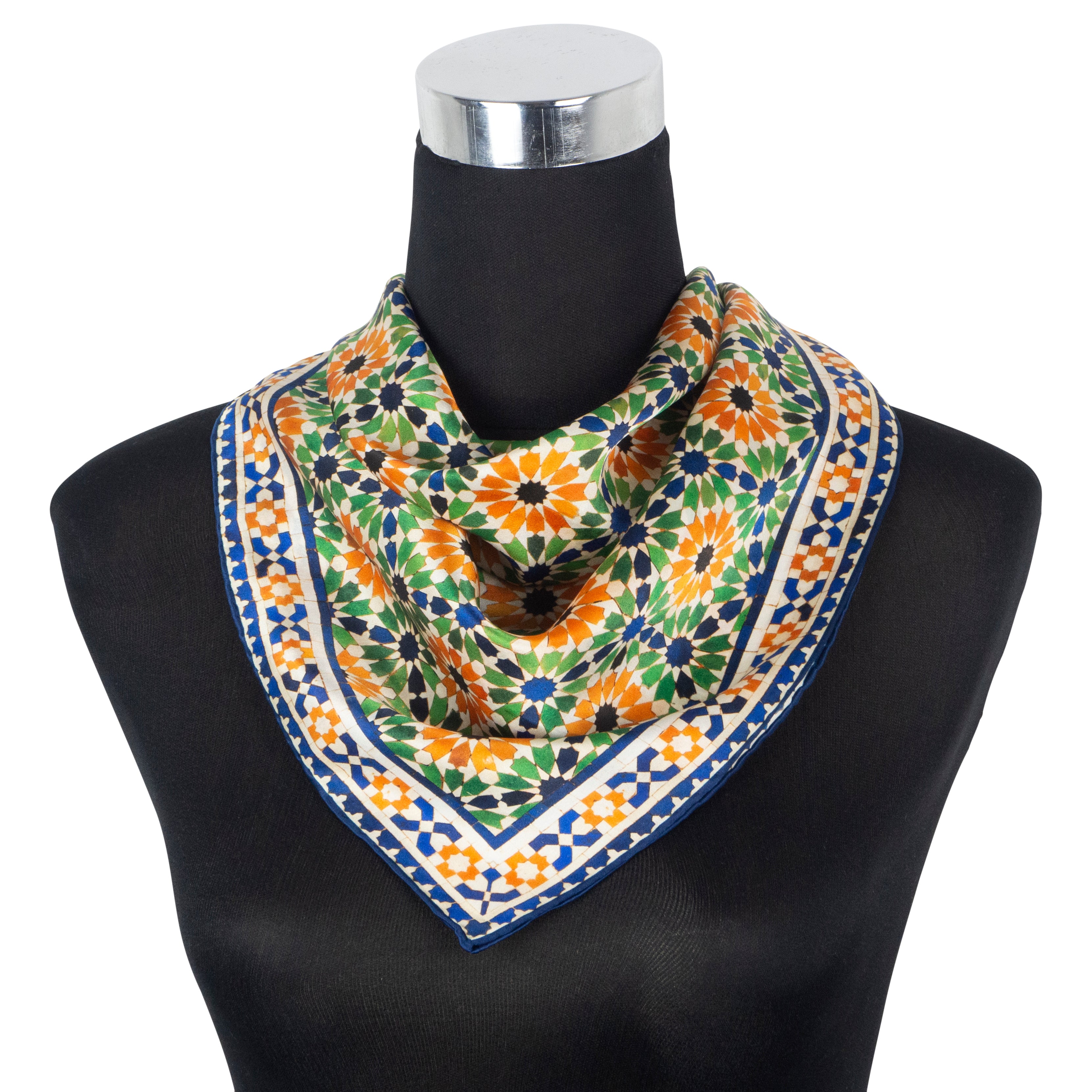 A mannequin displays the Multi Color Square Silk Scarf Nur, featuring a geometric floral pattern in orange, green, blue, and white, inspired by Islamic geometry, draped in a triangular style with hand rolled hems.