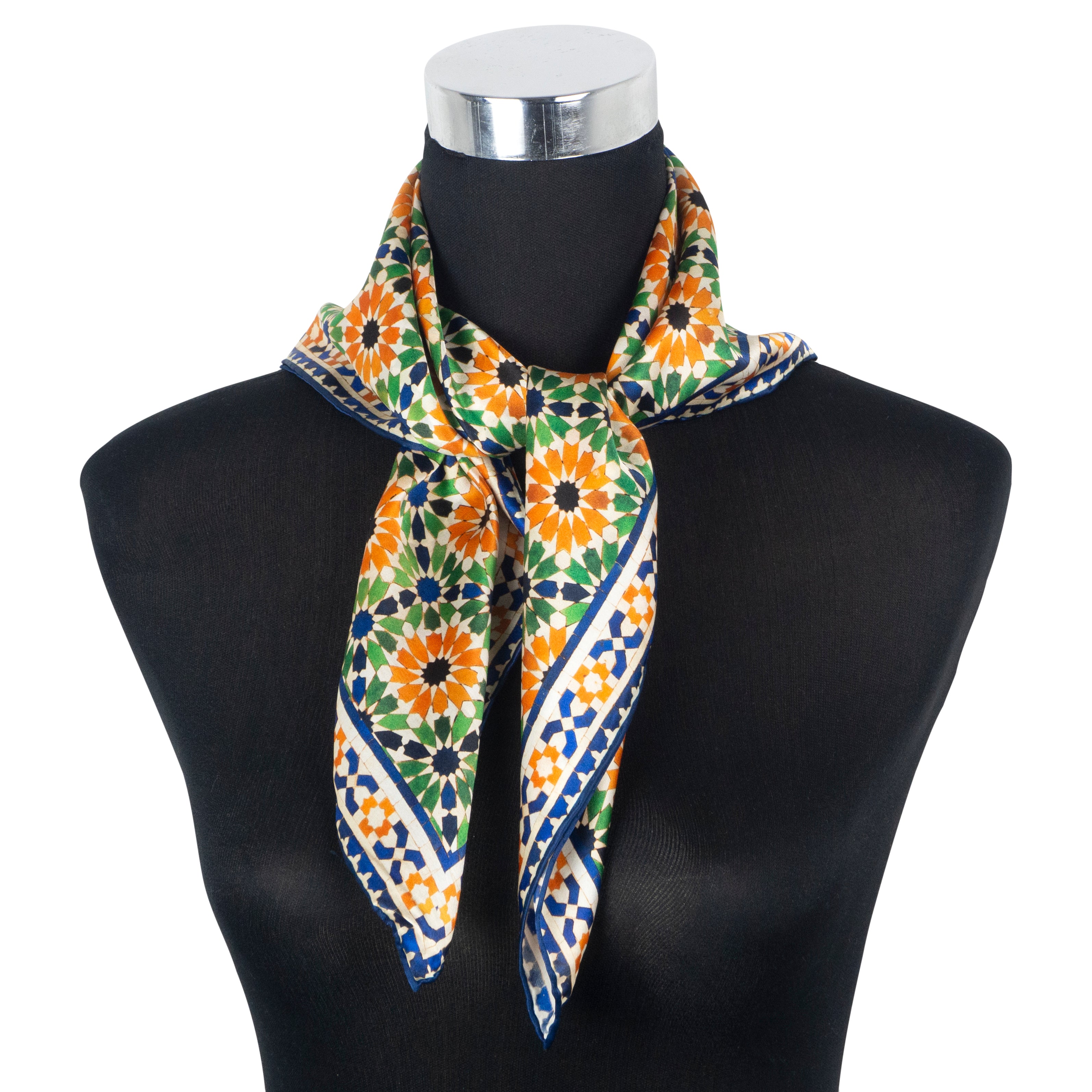 A mannequin displays the Multi Color Square Silk Scarf Nur, featuring a vibrant geometric floral pattern inspired by Islamic geometry, in orange, green, blue, and black, tied stylishly around its neck.