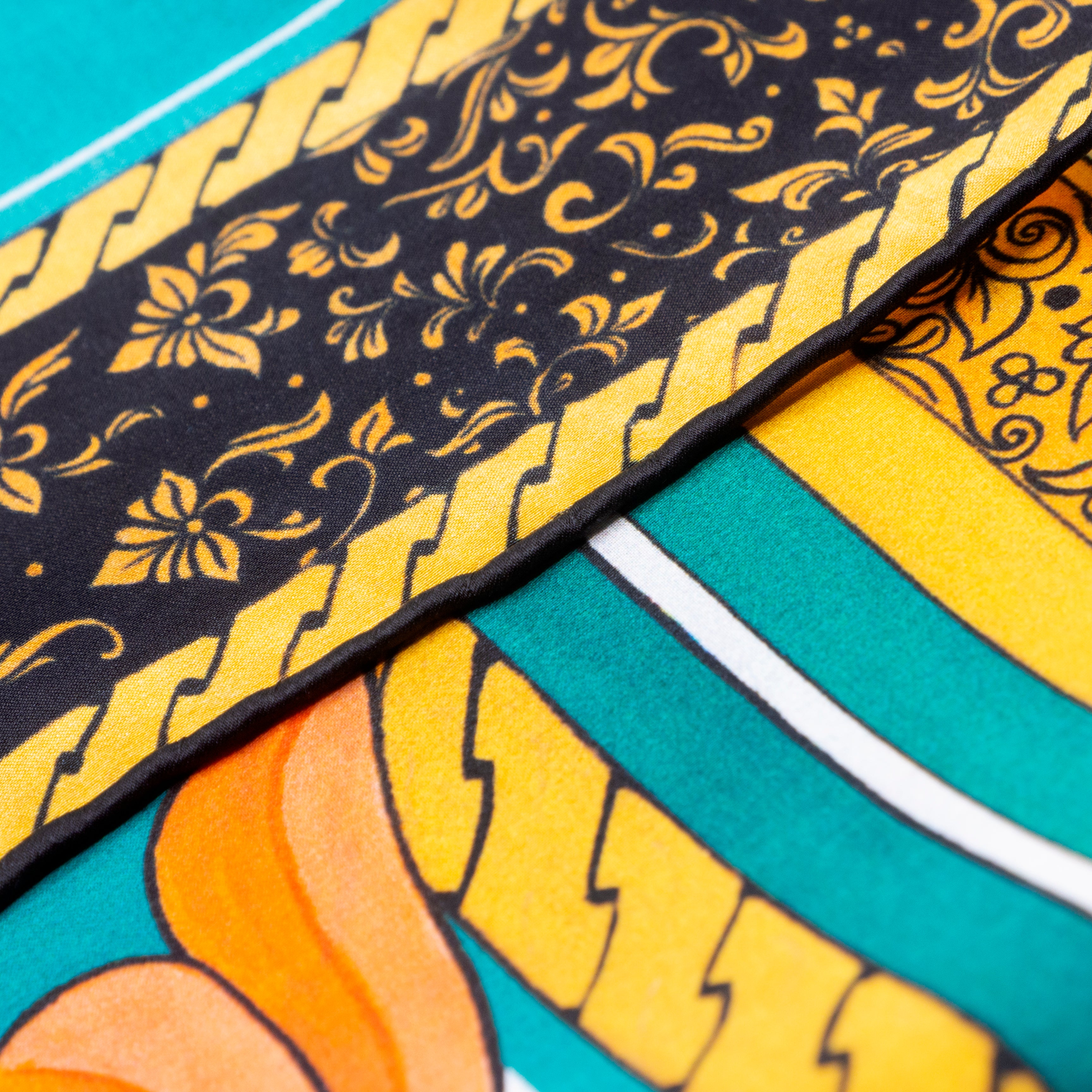 Close-up of the Floral Square Silk Scarf Olaya, showcasing intricate Islamic art-inspired patterns with black and yellow floral sections, yellow zigzag borders, orange accents, and green, white, and turquoise stripes with hand-rolled hems.