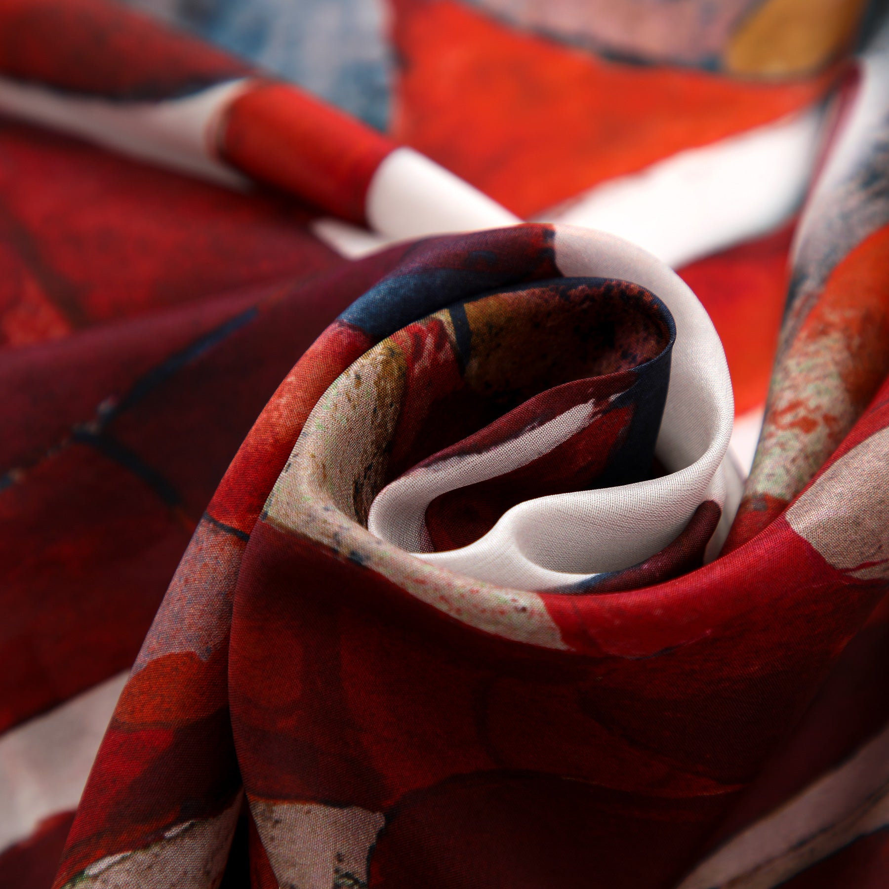 Close-up of the Navy Blue Silk Scarf Plumas al Viento, artistically twisted to showcase its smooth habotai silk texture and vibrant red, white, and navy abstract Mediterranean-inspired pattern.