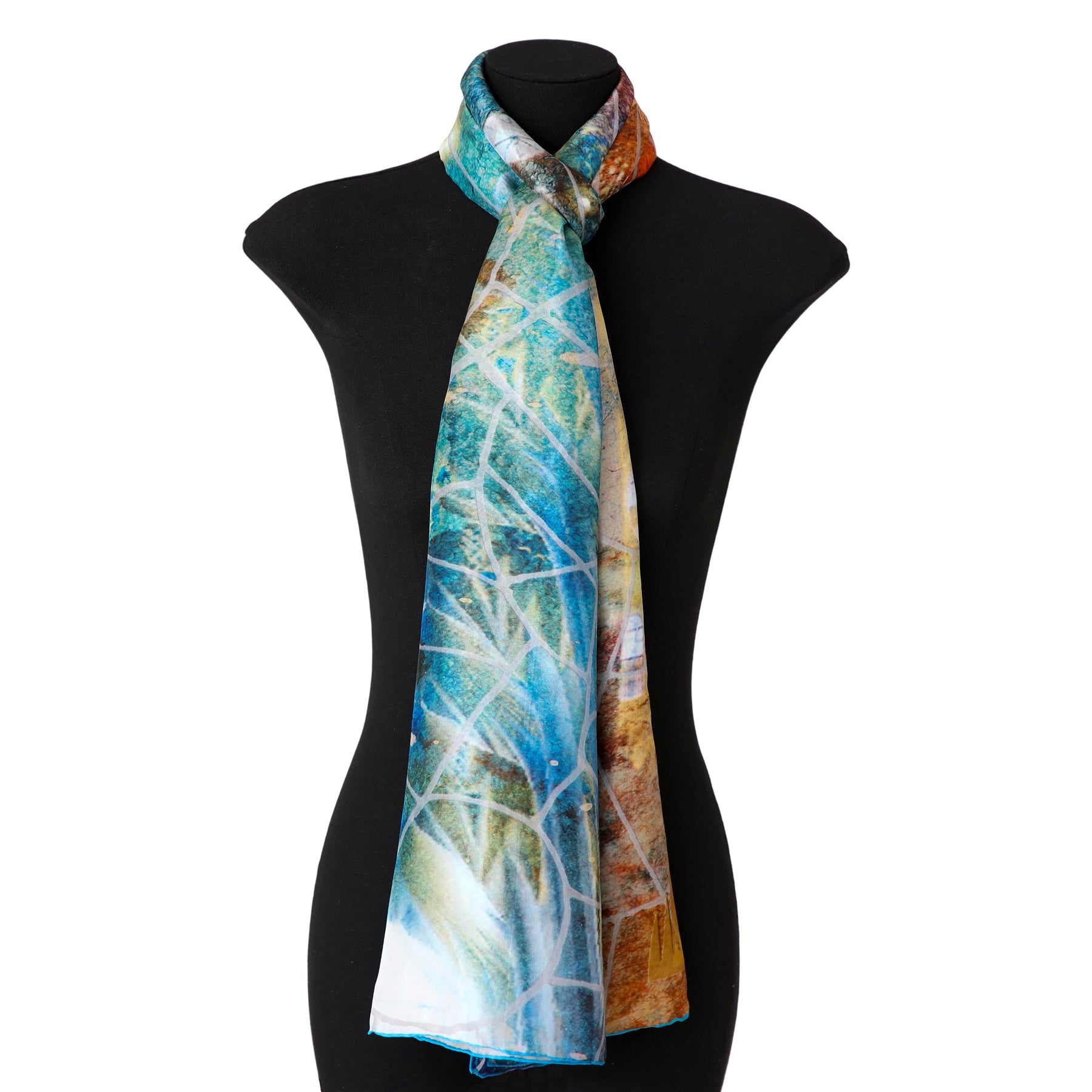 The Artisan Silk Scarf Estrellas del Templo features abstract Mediterranean patterns in blue, white, brown, and green, elegantly draped on a black mannequin—a stunning piece of wearable art.