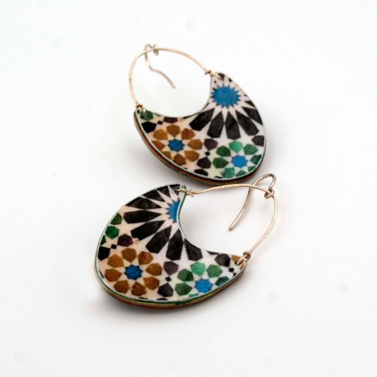 The Andalusian Tiles Inspired Earrings Almunecar by Azul Añil feature crescent shapes with blue, green, brown, and black mosaic patterns on white. Handmade in Spain, they echo traditional tile art with intricate geometric details and a delightful dangling hoop.