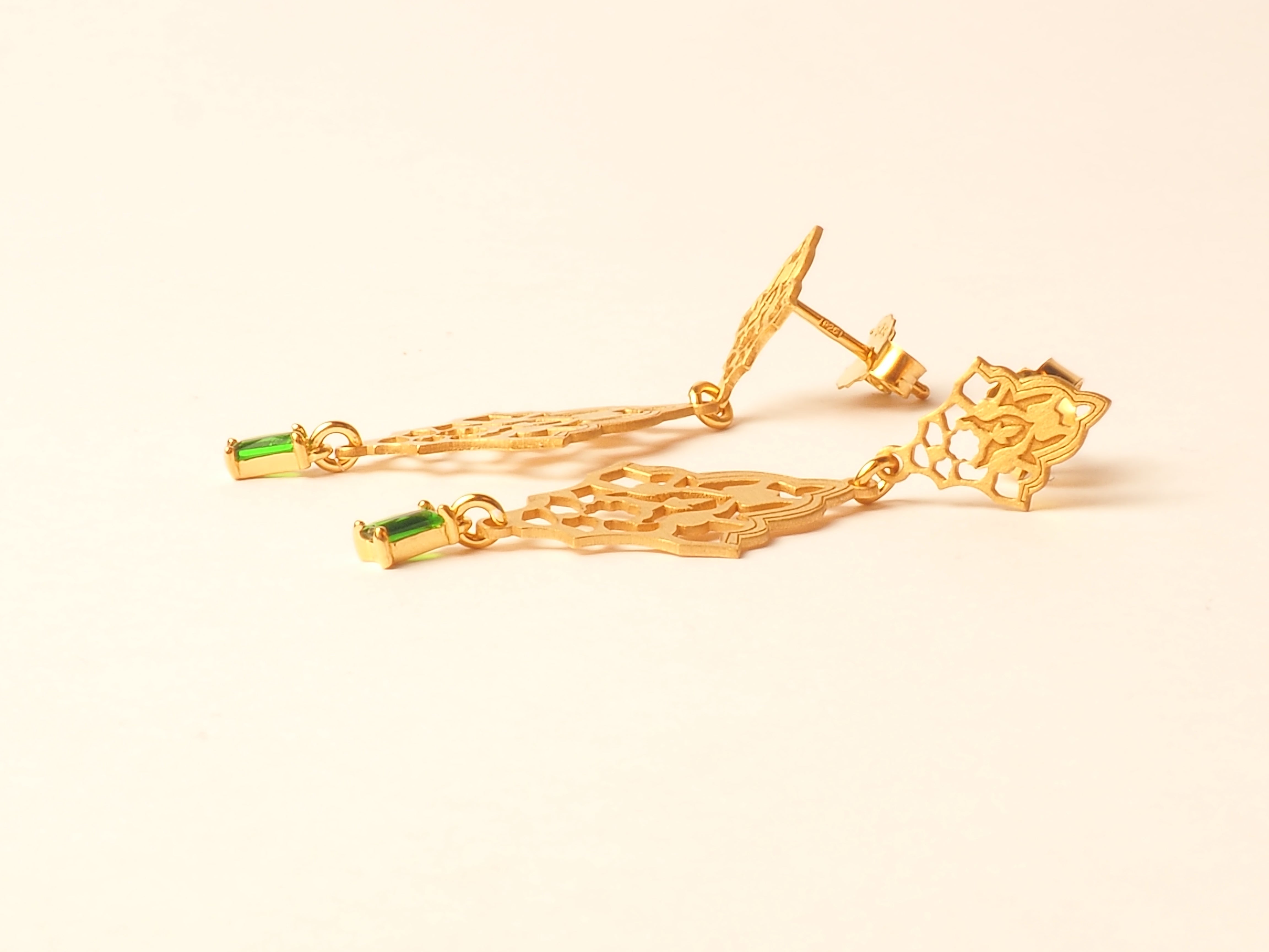 From Platónicas Sebka collection, the Double Emerald Gold Earrings take inspiration from Islamic art, showcasing intricate geometric designs with green rectangular gemstones. The elegant double-drop design includes a stud back and is beautifully displayed on a plain white background.