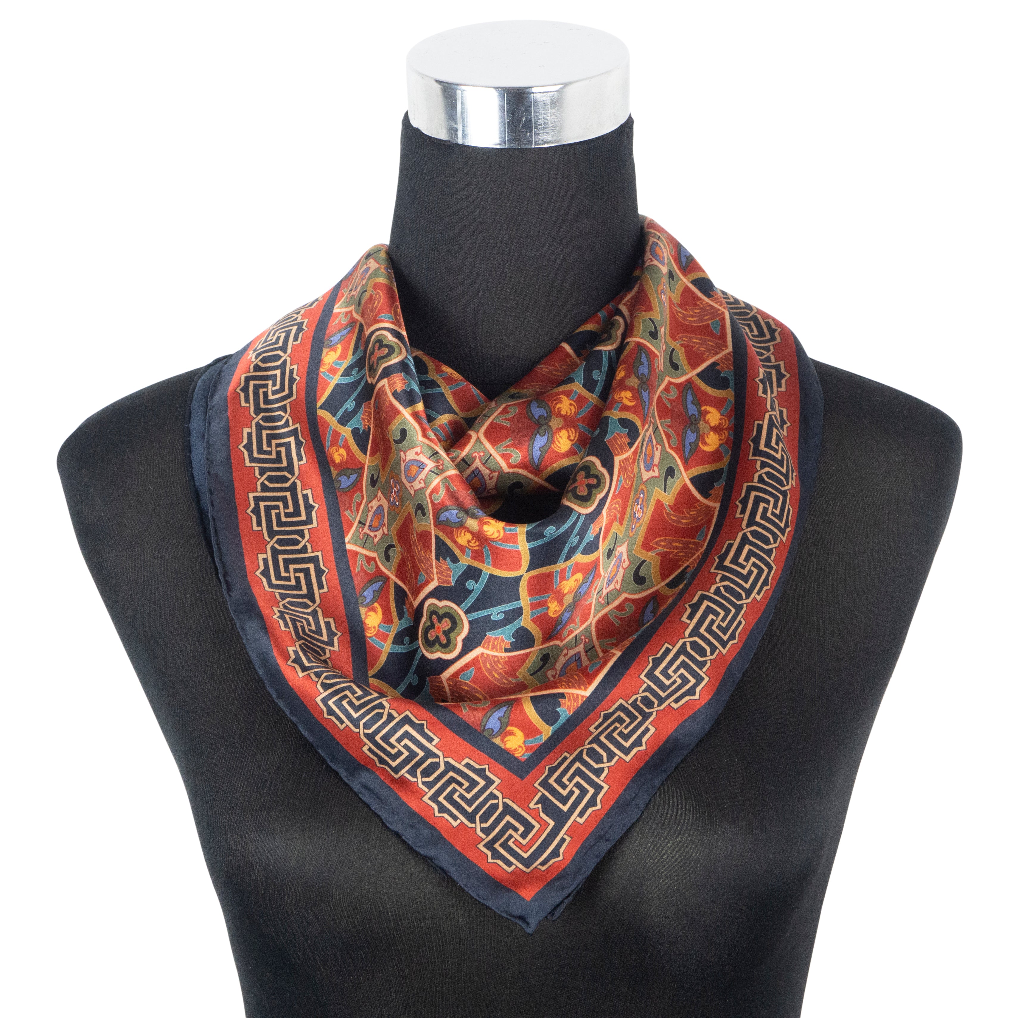 The Silk Square Neck Scarf Rania, with floral and geometric motifs inspired by Islamic art in red, orange, blue, and black tones and hand-rolled hems, is draped loosely around the mannequin’s neck.