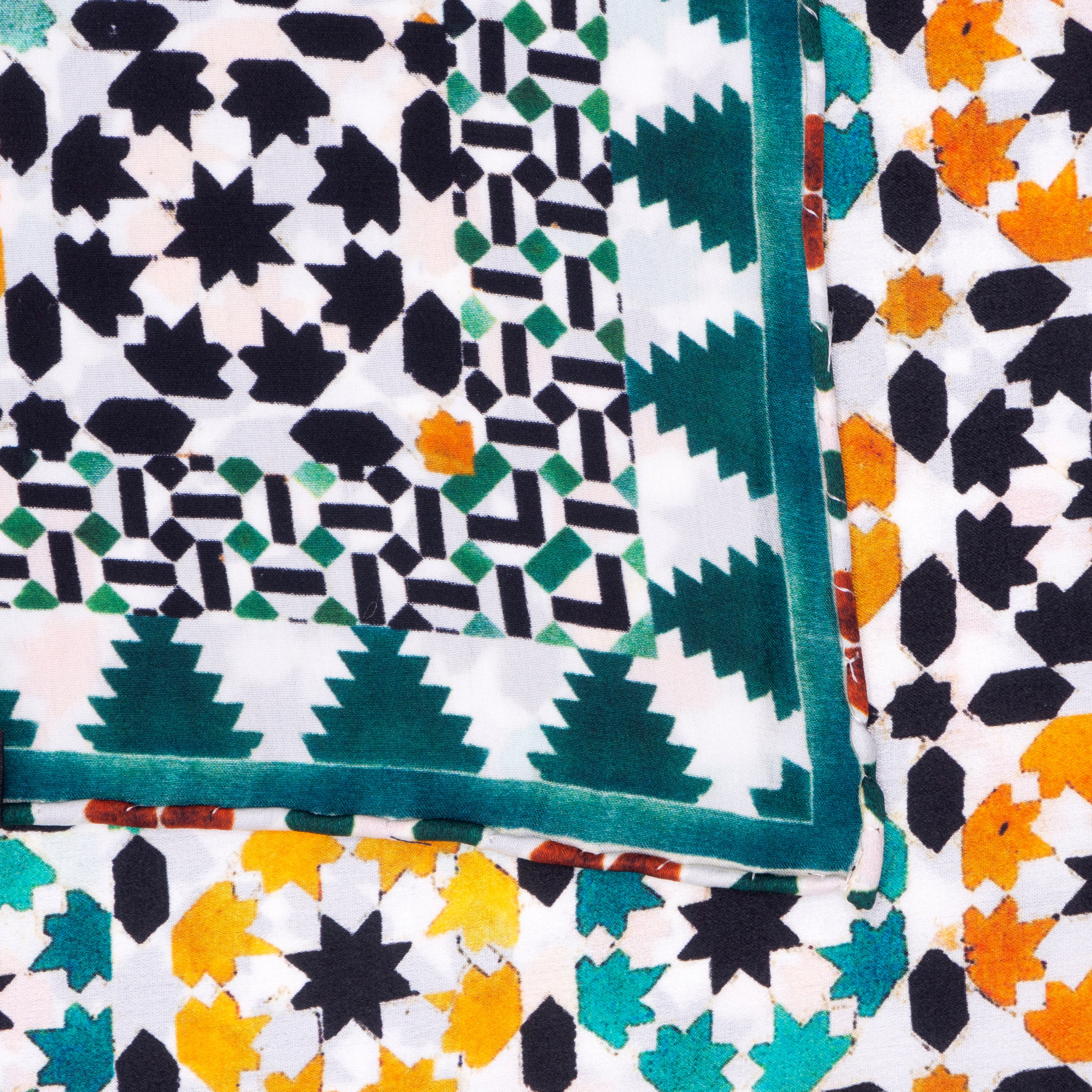 A close-up of overlapping fabrics with bold geometric art: the Islamic Art Square Silk Scarf Sabika Green features black, green, and white motifs with a border; another displays orange, teal, yellow, and black leaves on white.