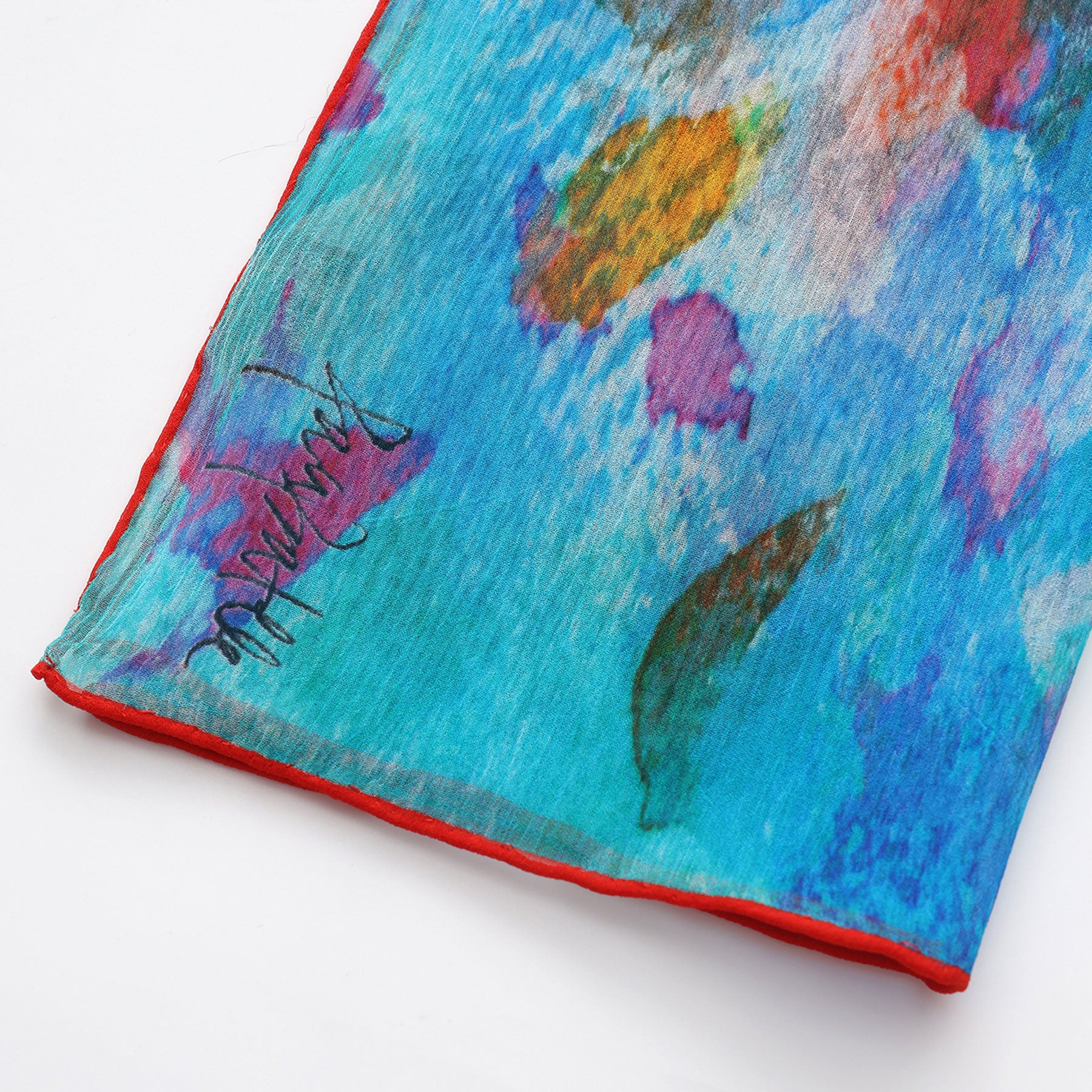 A close-up of the Multicolor Silk Scarf Mar Rojo in blue muslin silk, featuring abstract floral patterns, vivid red edges, and a black signature on the lower left—an elegant wearable artwork.