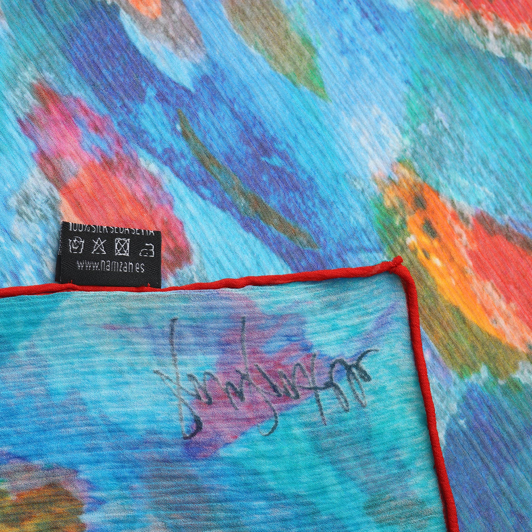 The Multicolor Silk Scarf Mar Rojo features abstract patterns in blue, green, red, and orange on muslin silk with a thin red border. A black tag with white care symbols is partially visible at the top left corner.