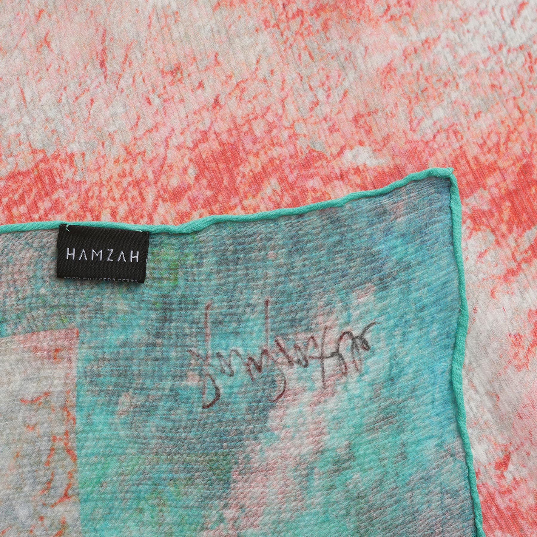 A close-up of the Multicolor Silk Scarf Fucsia Paradis in muslin silk, featuring a black HAMZAH label, green, blue, and red hues, a visible signature, and a finely stitched green border.