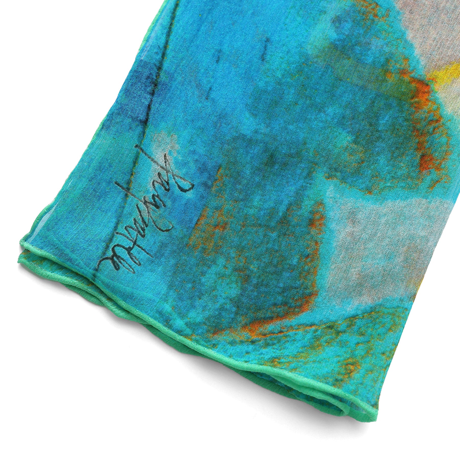 A close-up of the Multicolor Silk Scarf Oxido Marino reveals blue and green muslin silk with orange and yellow accents, a visible black signature near the edge, and a neatly stitched hem—wearable artwork in vibrant hues.