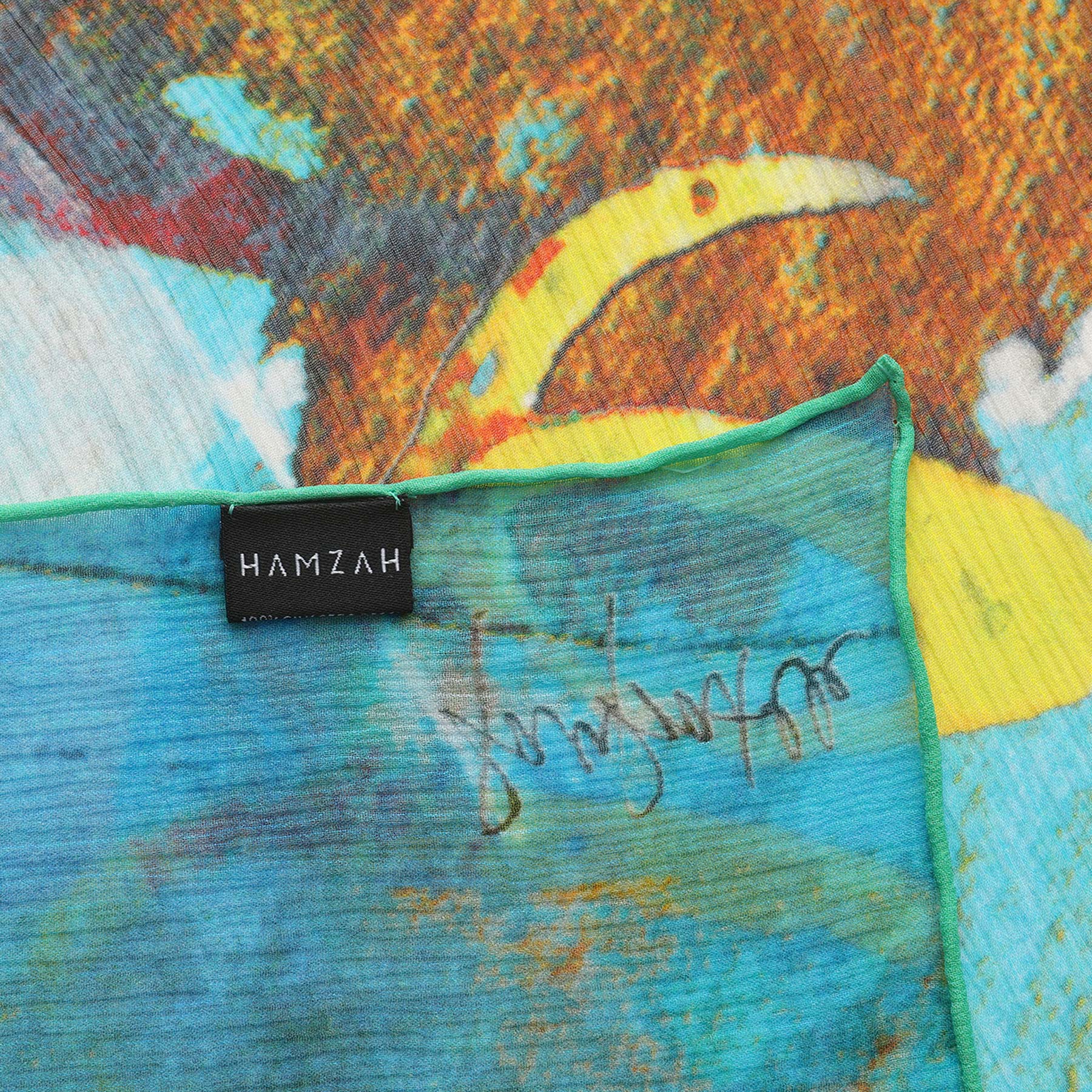 A close-up of the Multicolor Silk Scarf Oxido Marino reveals its vibrant abstract pattern, black HAMZAH label, green-stitched hem, and an artistic handwritten signature—capturing the essence of wearable art.