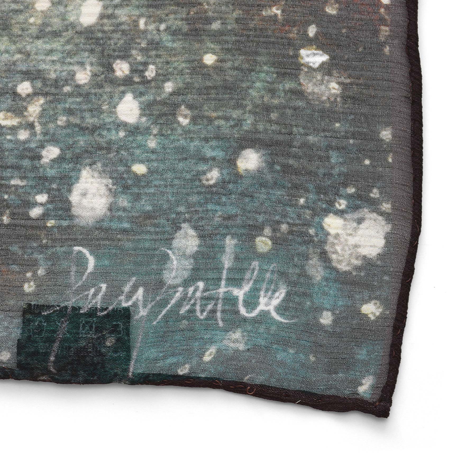 A close-up of the Multicolor Silk Scarf Polvo Cósmico reveals a speckled abstract pattern with white spots on a dark background, a white signature detail along the edge, and a dark finished border—making it wearable artwork.