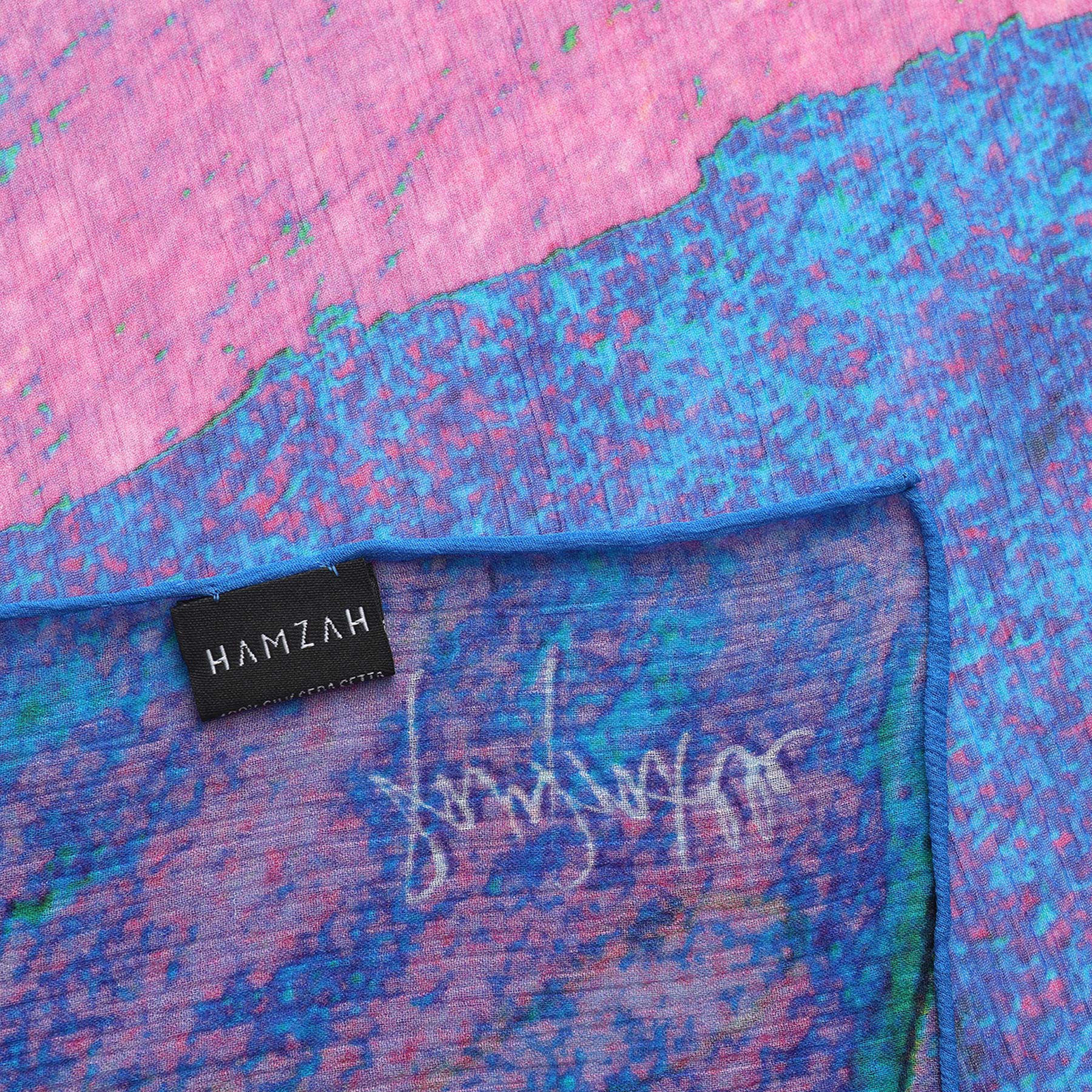 A close-up of the Multicolor Silk Scarf Océano Fucsia, featuring vibrant blue, pink, and purple abstract patterns with a black HAMZAH label and signature-style design—perfect as wearable art.