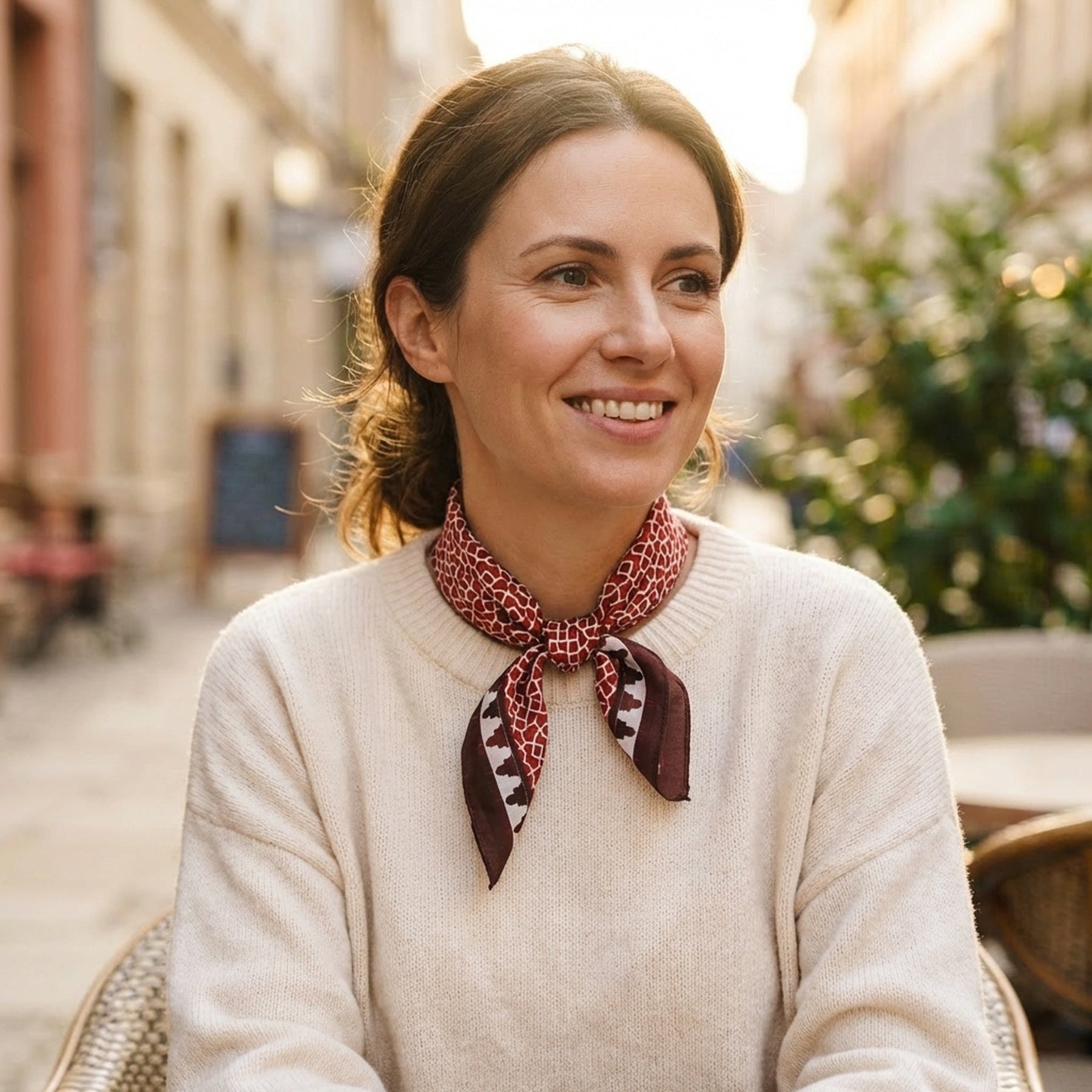 A woman with brown hair tied back, wearing a cream sweater and a Red Square Scarf 100% Silk, smiles while sitting at an outdoor café on a sunny street.