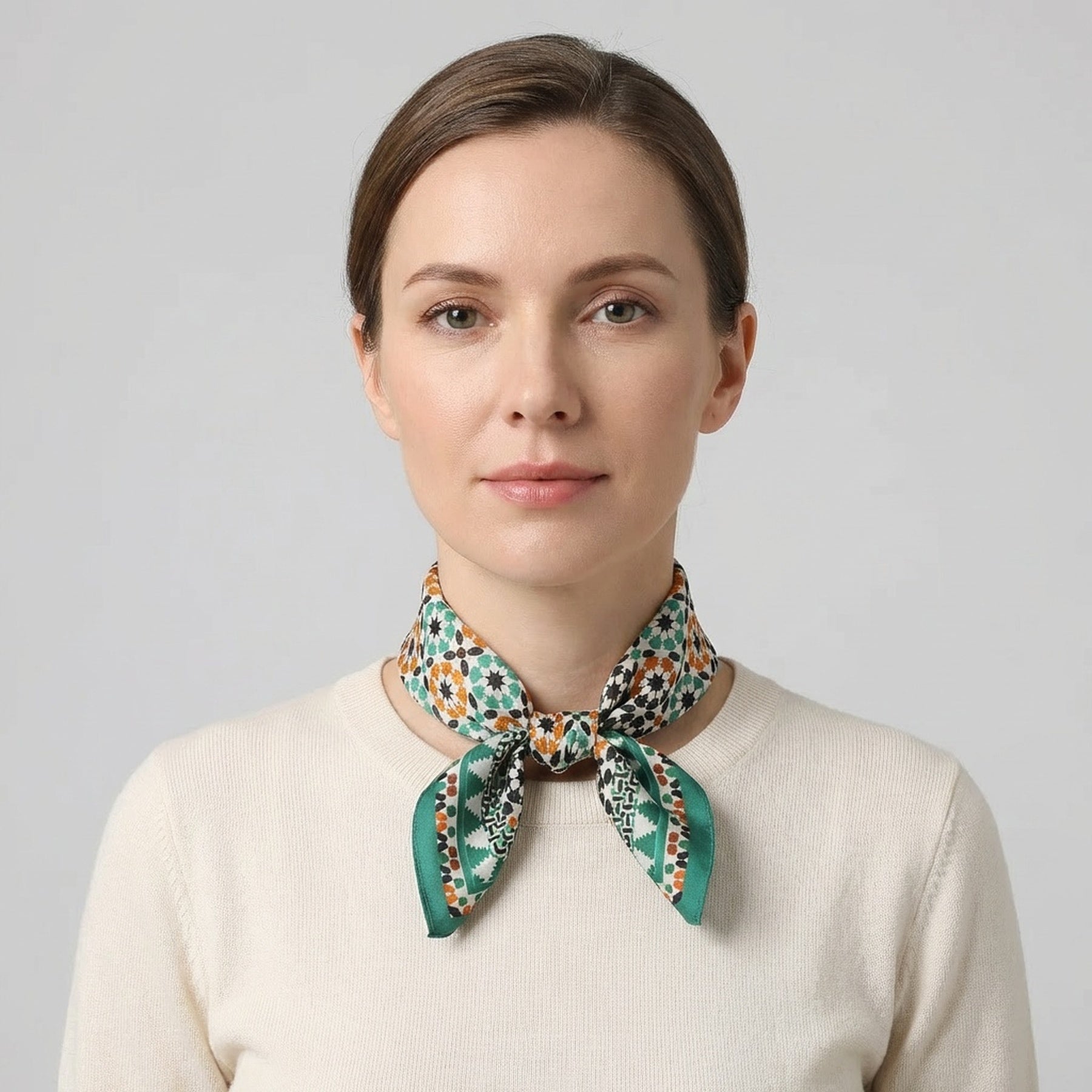 A woman with light skin and brown hair tied back wears a cream sweater and the Green Square Silk Scarf Sabika—brown with green and orange accents—tied around her neck. She faces forward against a plain background with a neutral expression.