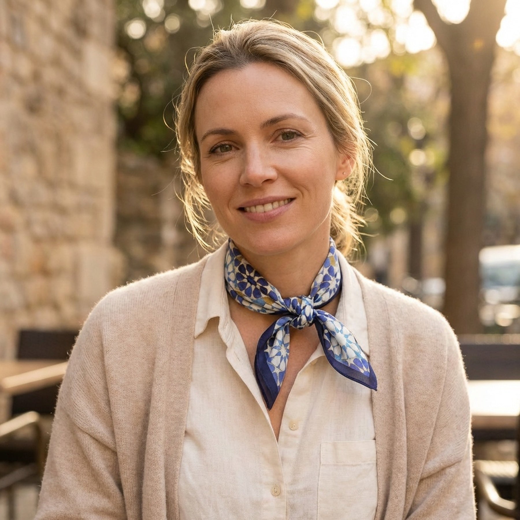 A blonde woman smiles outdoors, wearing a beige cardigan, white shirt, and an elegant Blue Neck Scarf 100% Silk around her neck. Sunlight filters through trees and stone buildings in the background.