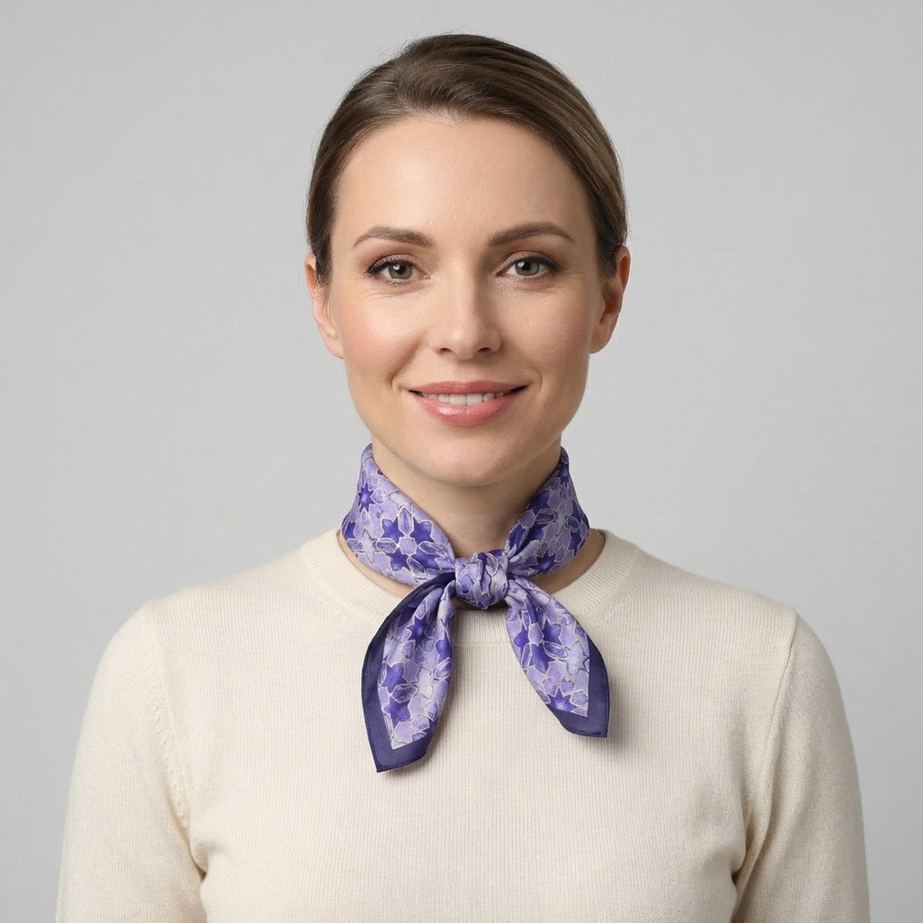 A woman with light brown hair tied back, wearing a cream-colored top and the Square Silk Scarf for Women Purple, featuring an Islamic art print, smiles at the camera against a plain gray background.