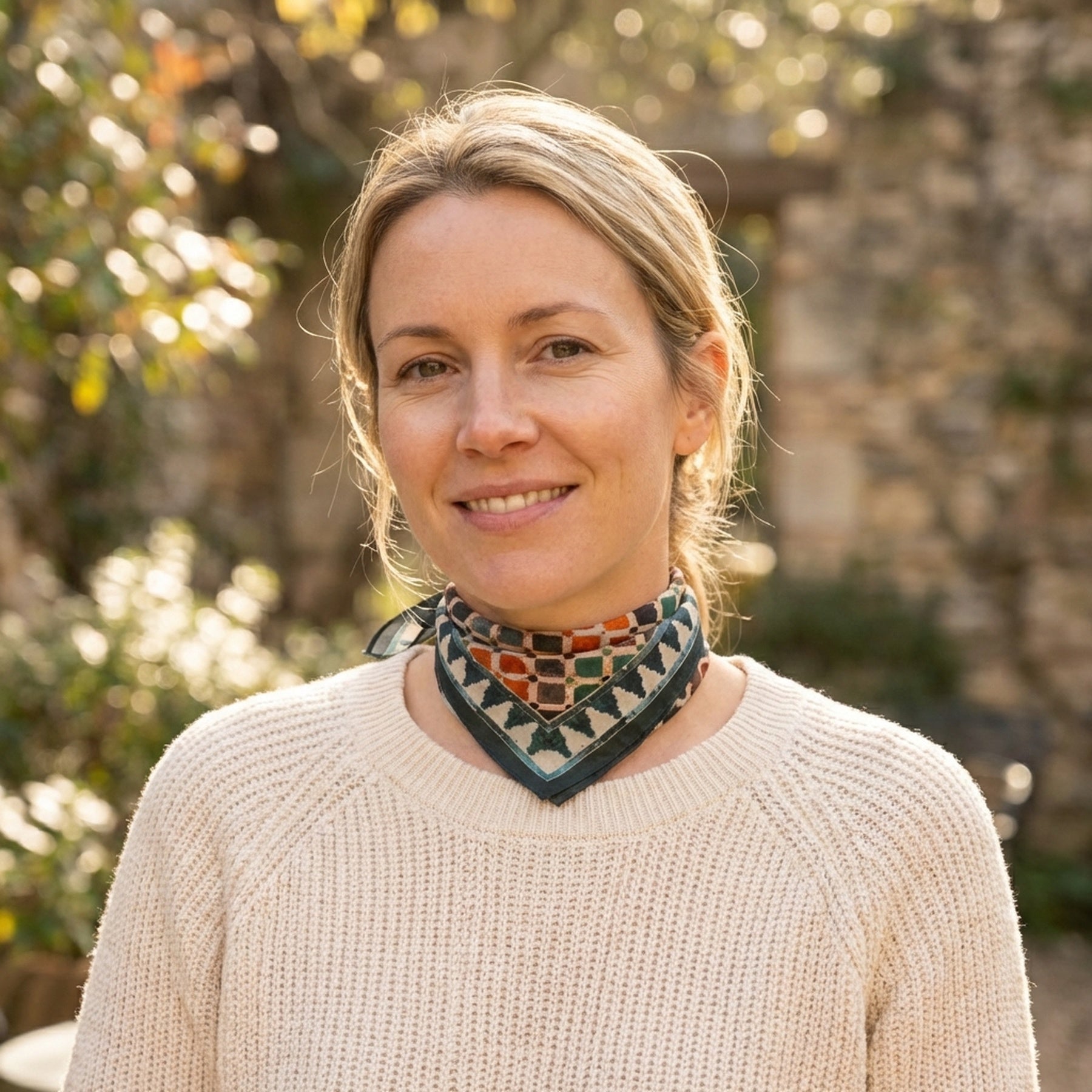 A woman with blonde hair tied back, wearing a cream sweater and the Colorful Square Scarf Comares, smiles softly outdoors in natural sunlight with a blurred background of greenery and stone.