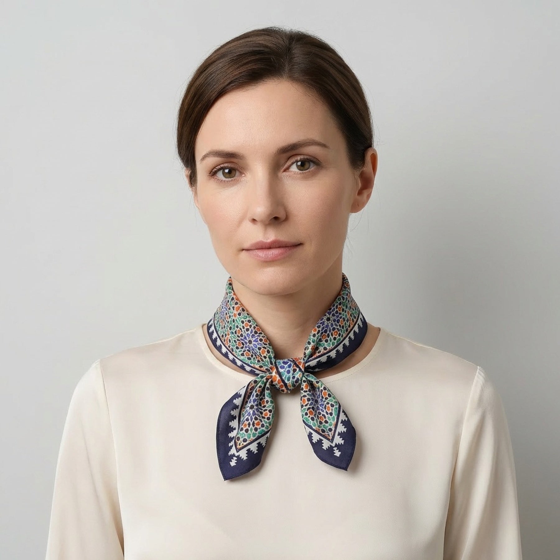 A woman with brown hair in a low bun wears a cream blouse and the Square Scarf Blue Madiya—a navy blue square scarf with a colorful floral pattern—tied around her neck, standing against a light gray background.