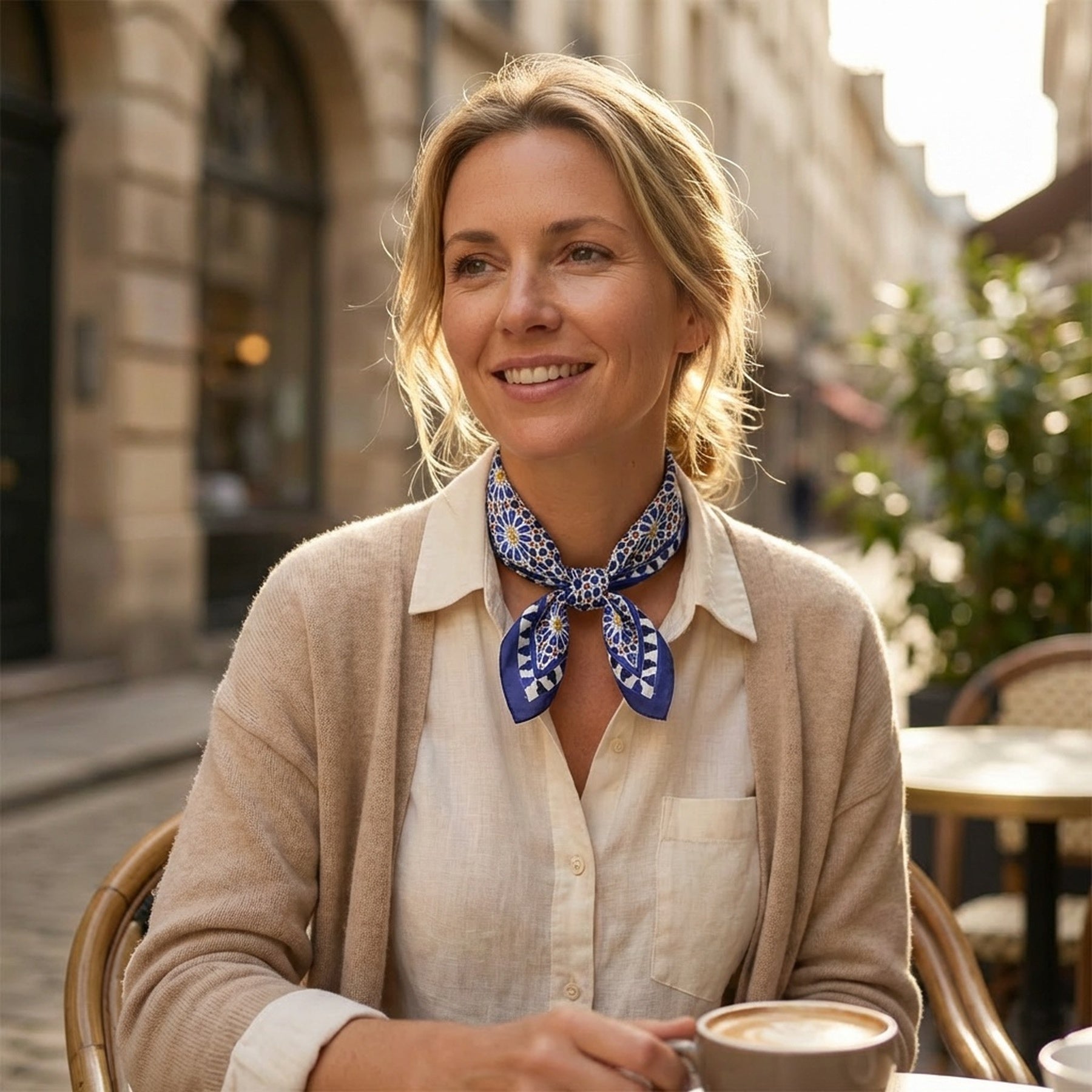 Wearing the Blue Square Scarf Zellige v2 and a beige cardigan, a woman smiles at an outdoor café table with coffee, set against a sunlit street of historic buildings.